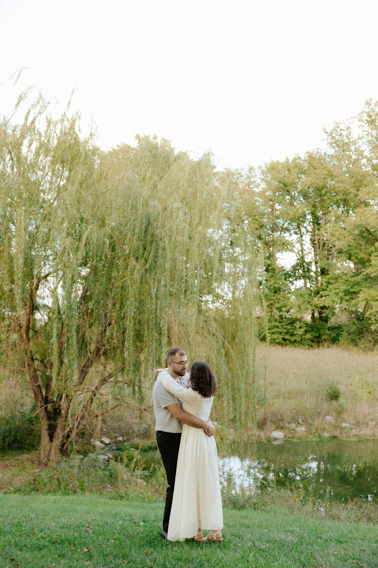 A couple embraces under a weeping willow tree by a tranquil pond in a romantic outdoor setting.
