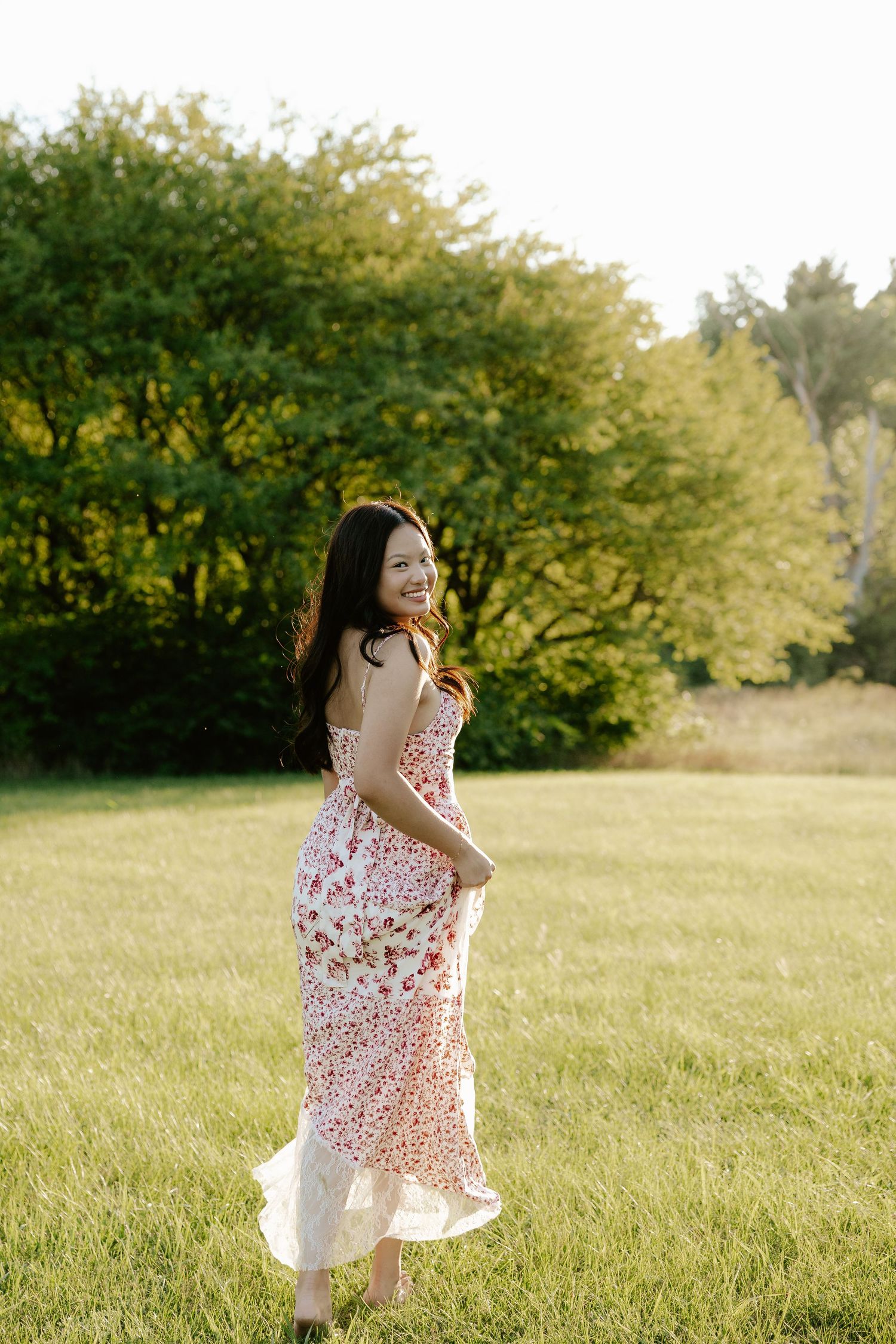 A girl in a white floral dress twirls playfully in a sunlit meadow surrounded by green trees at Pioneers Park in Lincoln Nebraska.
