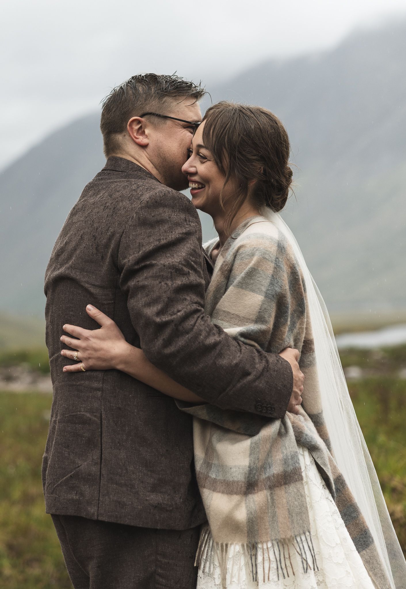 Couple embracing and smiling in wedding attire with misty mountains backdrop, bride in plaid shawl and veil.