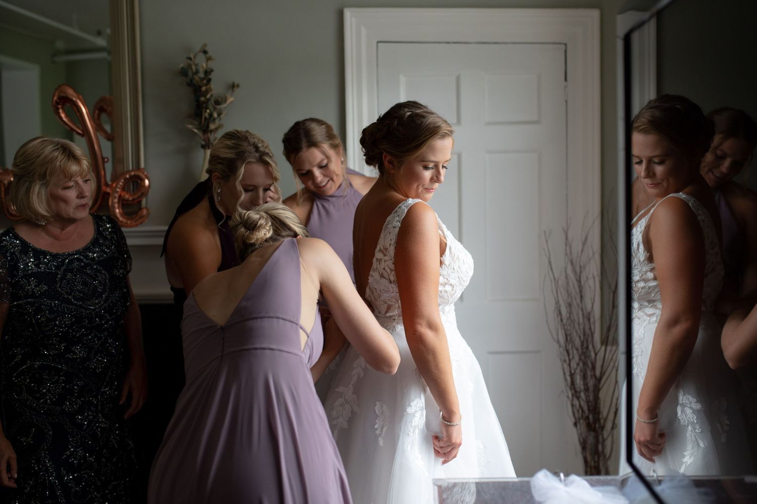A bridal party gets ready in a sunlit room as bridesmaids in lavender dresses help with final preparations.