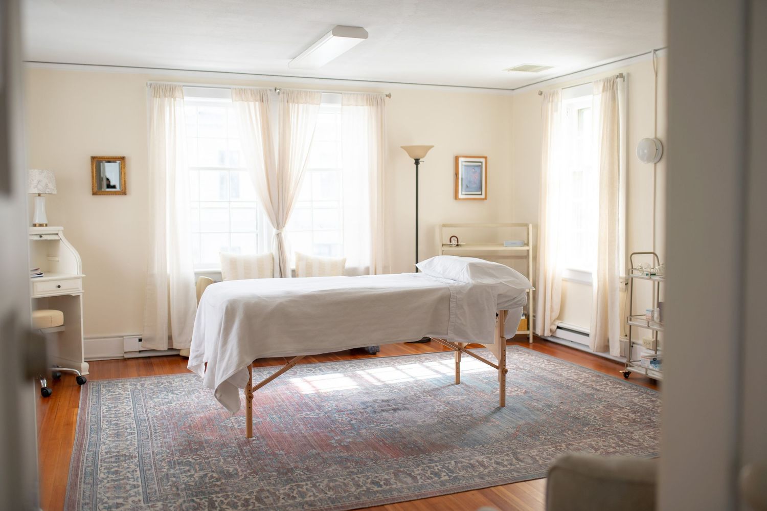 A bright bedroom with hardwood floors features a simple metal bed, ornate rug, and sunlit windows with sheer curtains.