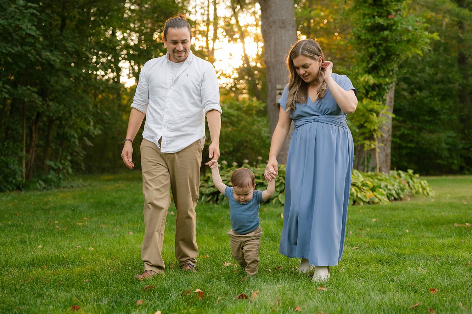 A family walks together on a green lawn at sunset, with trees and garden foliage in the background.