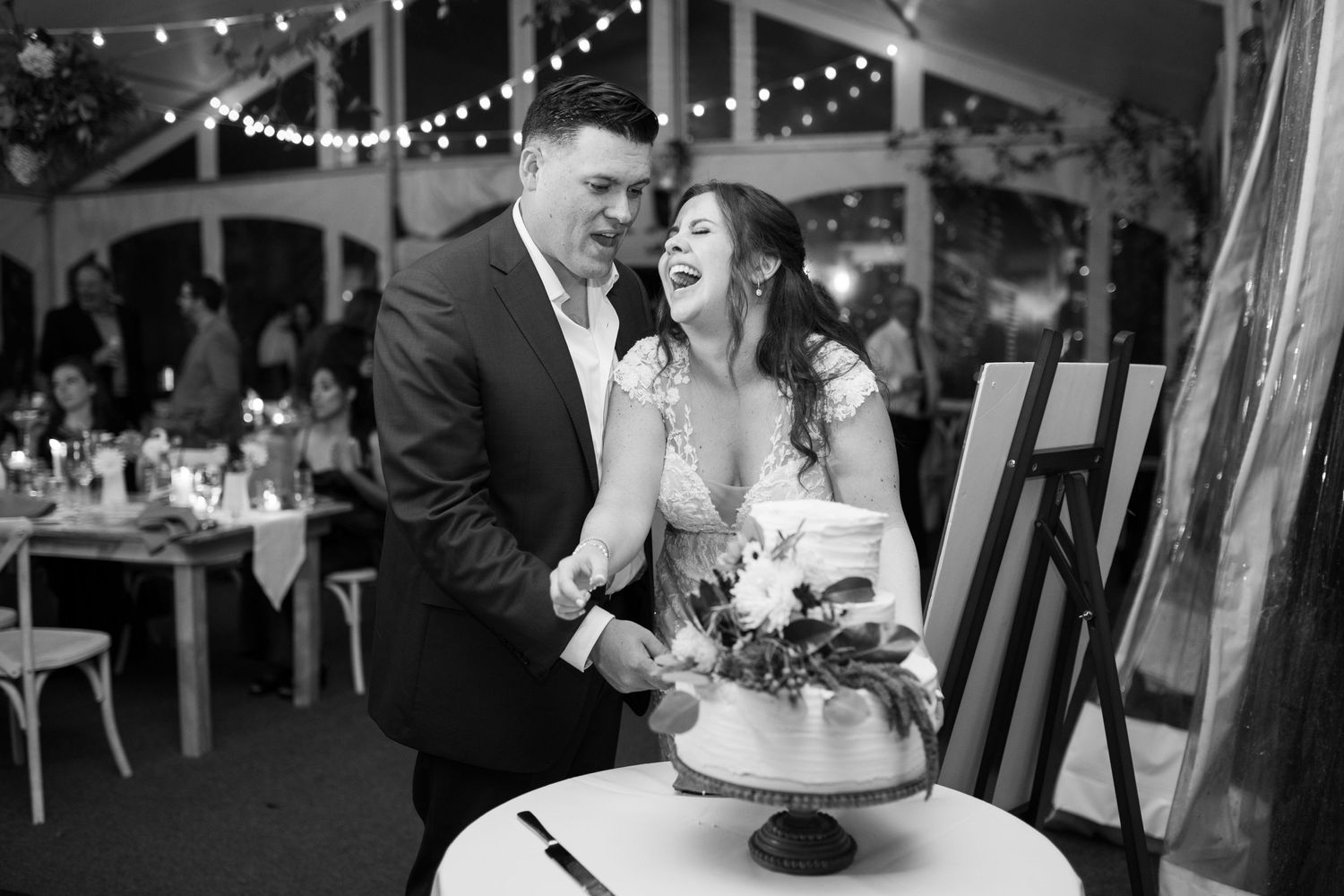 A black and white photo of newlyweds laughing while cutting their wedding cake.