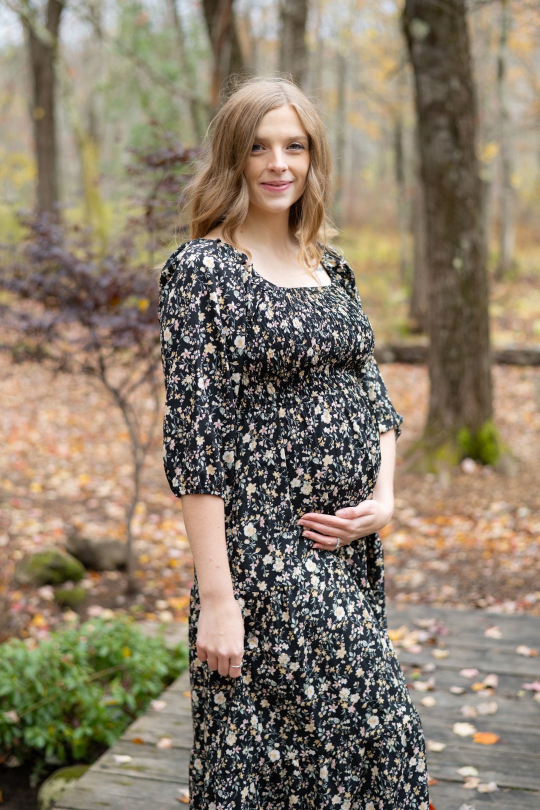 A pregnant woman in a floral print dress stands on a wooden walkway surrounded by autumn foliage in the woods.