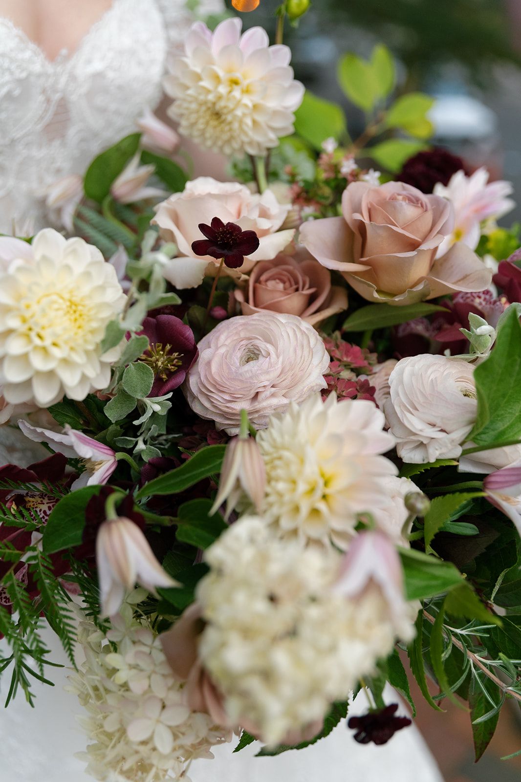 Close-up of romantic wedding bouquet with blush roses, white dahlias, and ranunculus flowers against soft green foliage.