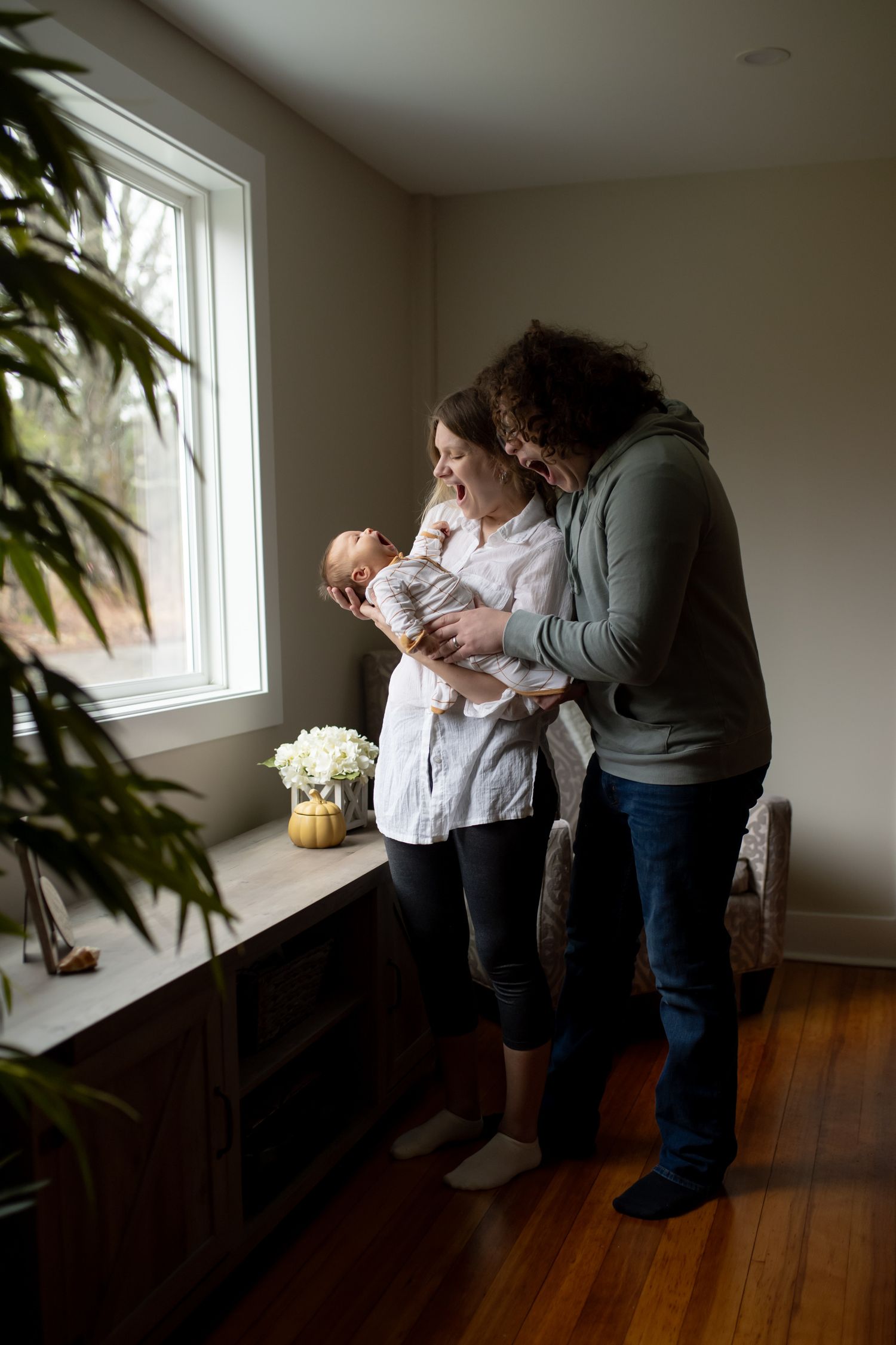 Two people stand in a sunlit room with hardwood floors and white walls near a window, creating a tender moment.