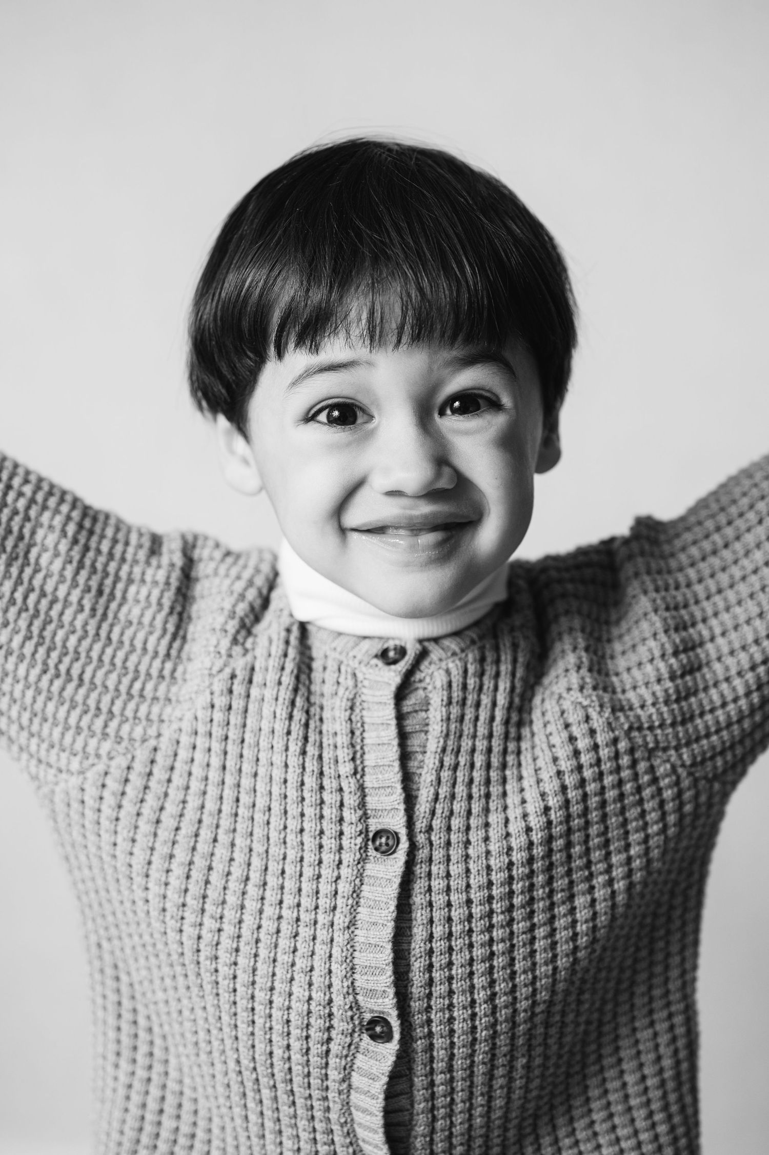 Black and white portrait of a child wearing a knit cardigan sweater with a joyful expression and arms raised.