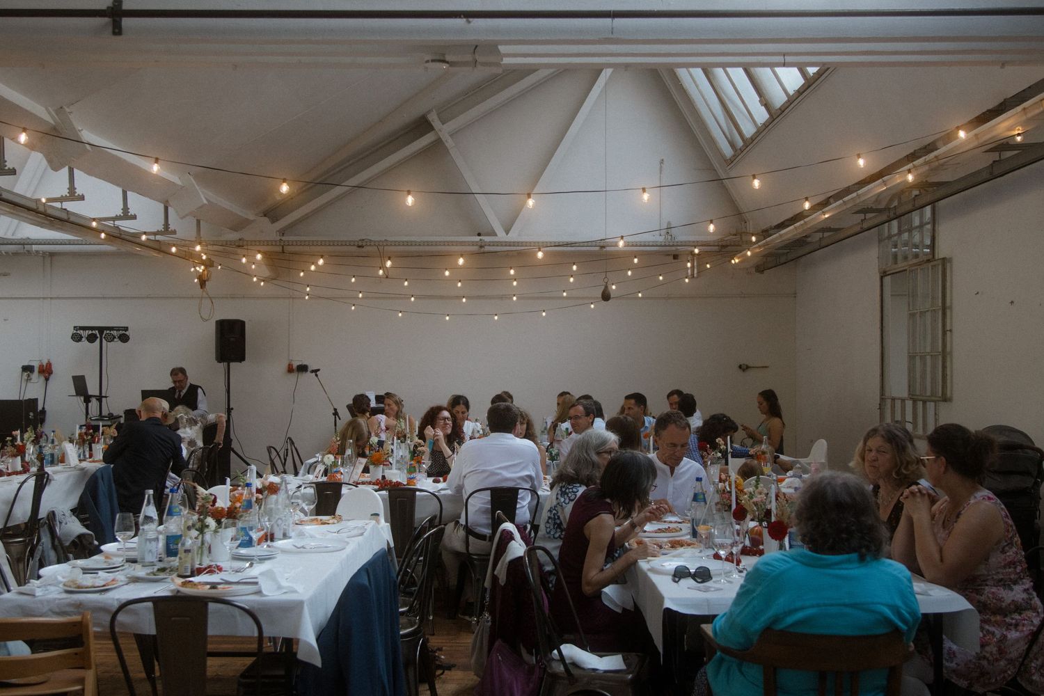 Wedding reception guests sit at round tables decorated in white linens inside a bright, airy venue with skylights.