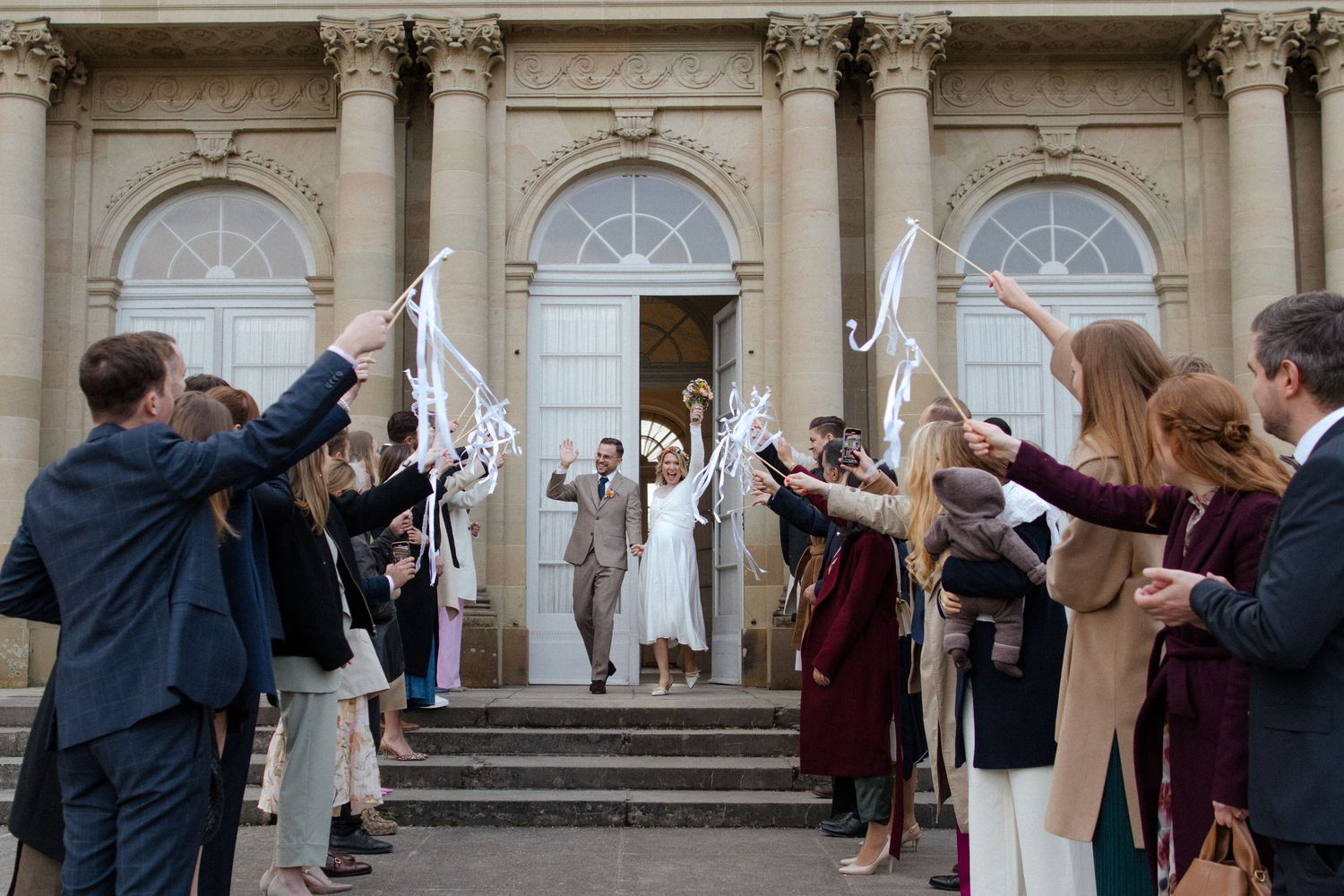 A crowd gathers for a wedding ceremony at elegant castle steps with stone archways and classical architecture.