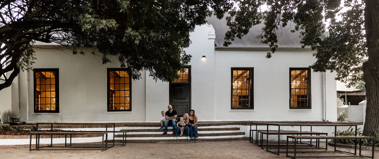 A glowing white colonial-style building at dusk with illuminated windows and people seated on benches outside.