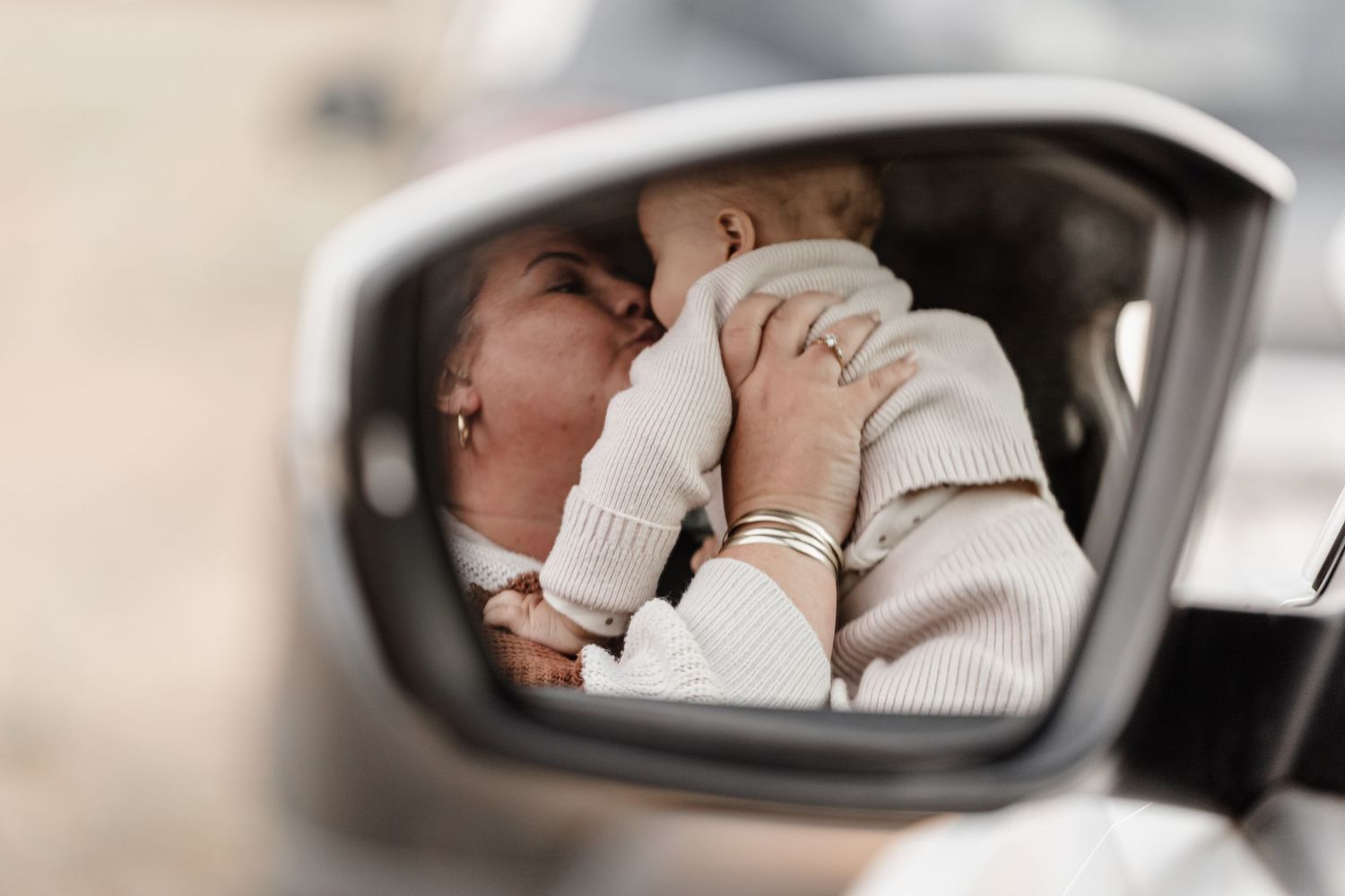 Side mirror reflection shows car passenger in cream sweater covering their face with hands while seated.