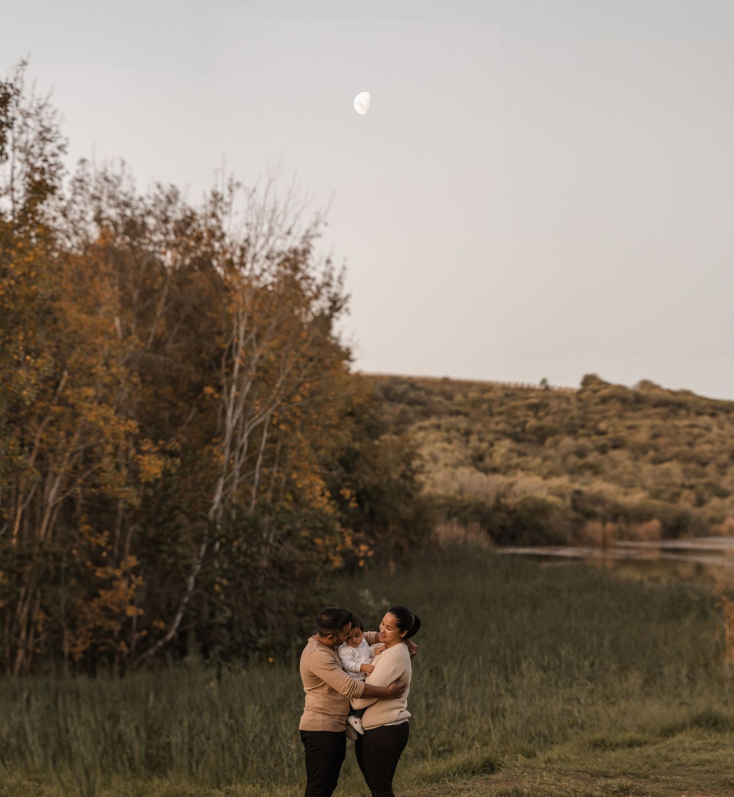 A romantic couple embraces in a serene landscape with autumn trees and a soft moon in the hazy sky.