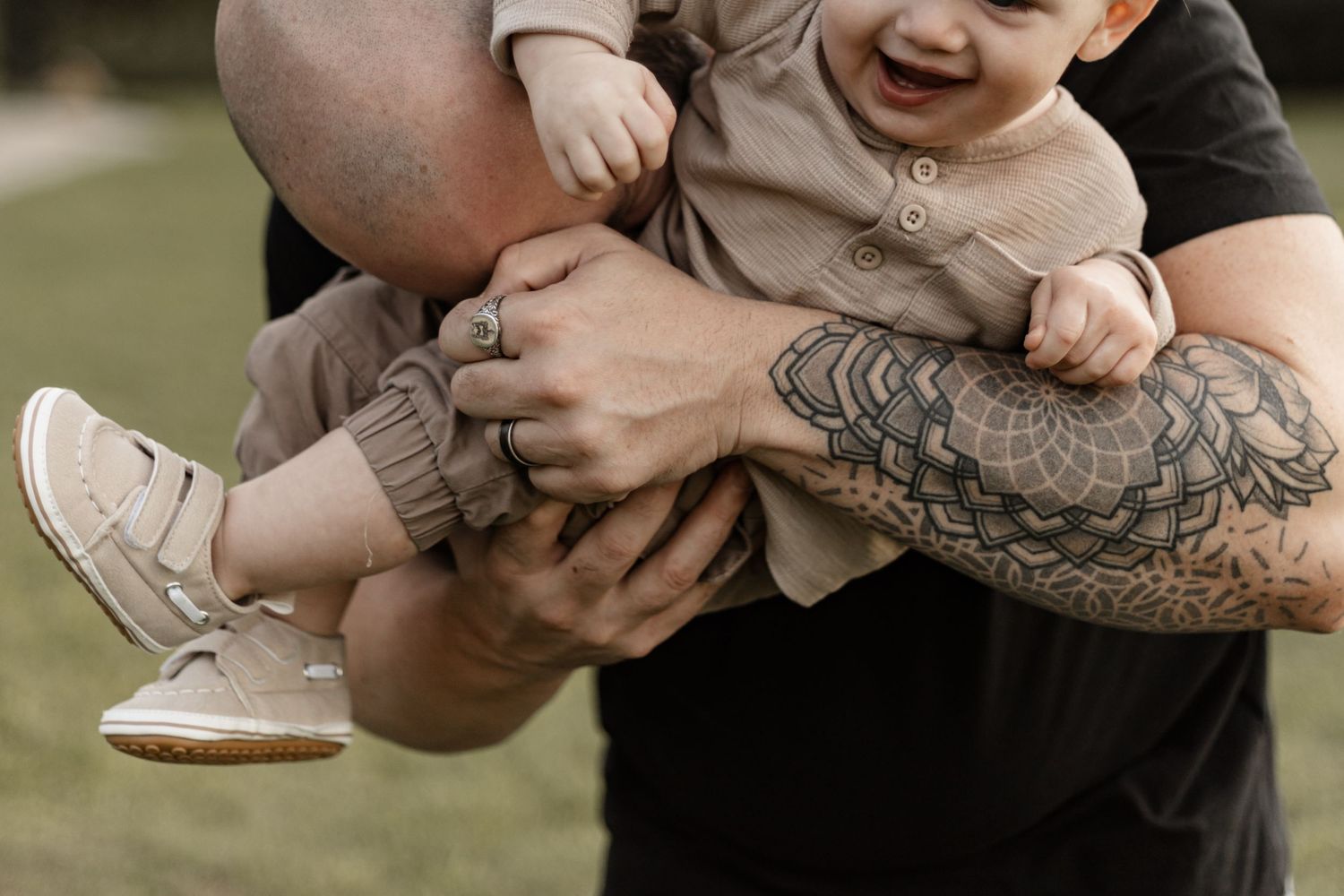 Close-up view of arms with sleeve tattoos holding a laughing baby wearing cream-colored clothing and white shoes.