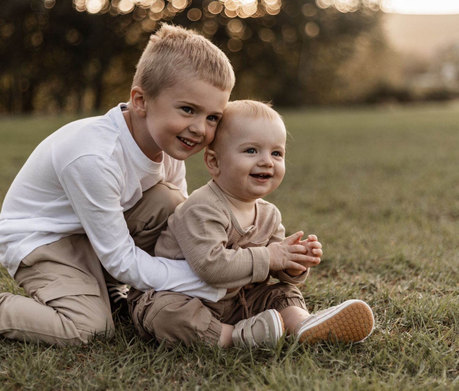 Two young children in coordinated neutral outfits sit together smiling on grass during golden hour sunset.