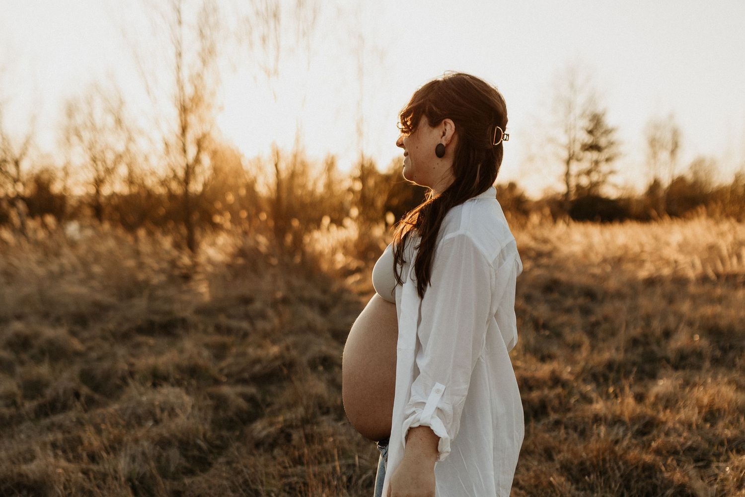 Pregnant woman in white shirt standing in golden field at sunset, cradling baby bump in serene maternity photo.