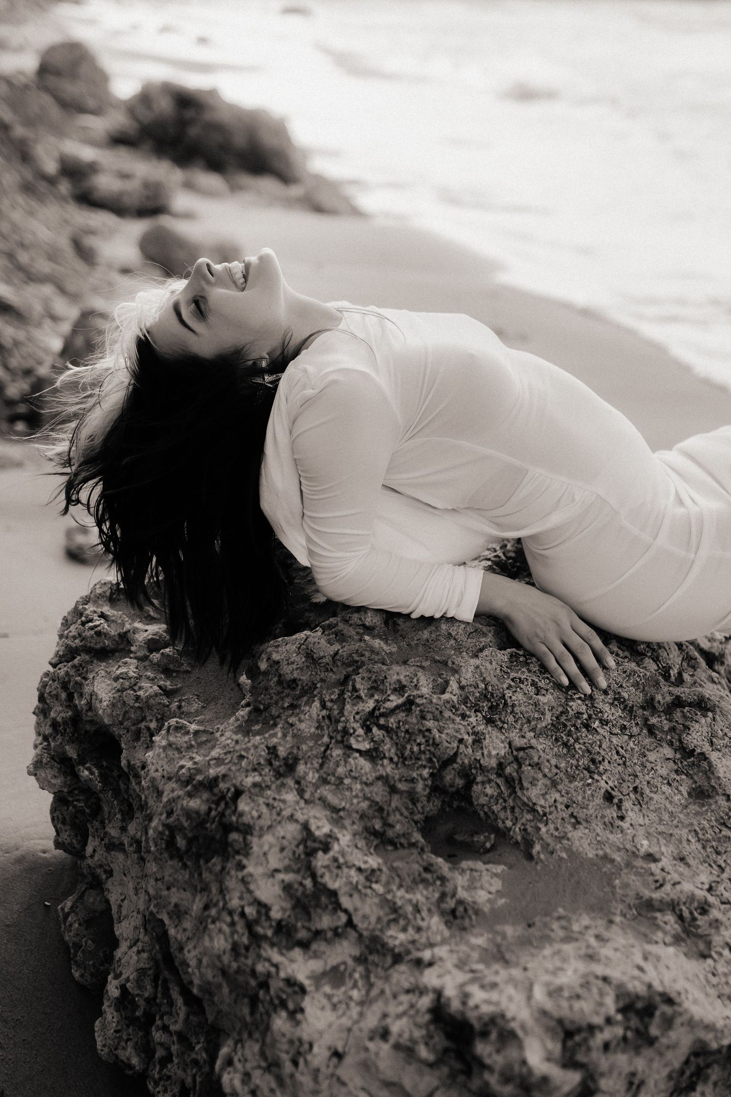 Black and white photo of a woman in white dress reclining on a rock by the ocean beach.