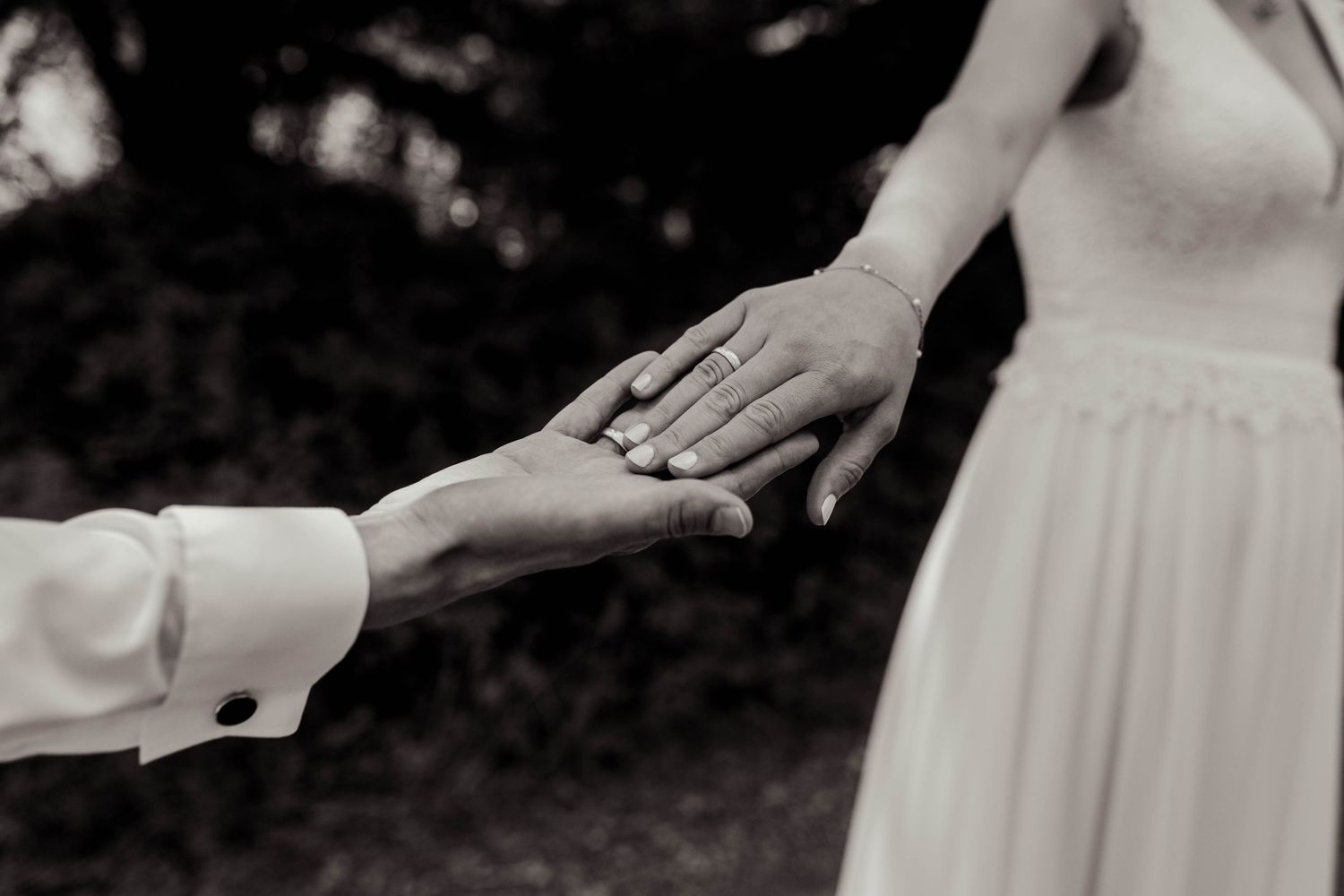 Black and white photo of bride and groom holding hands, showcasing wedding ring during romantic outdoor ceremony.