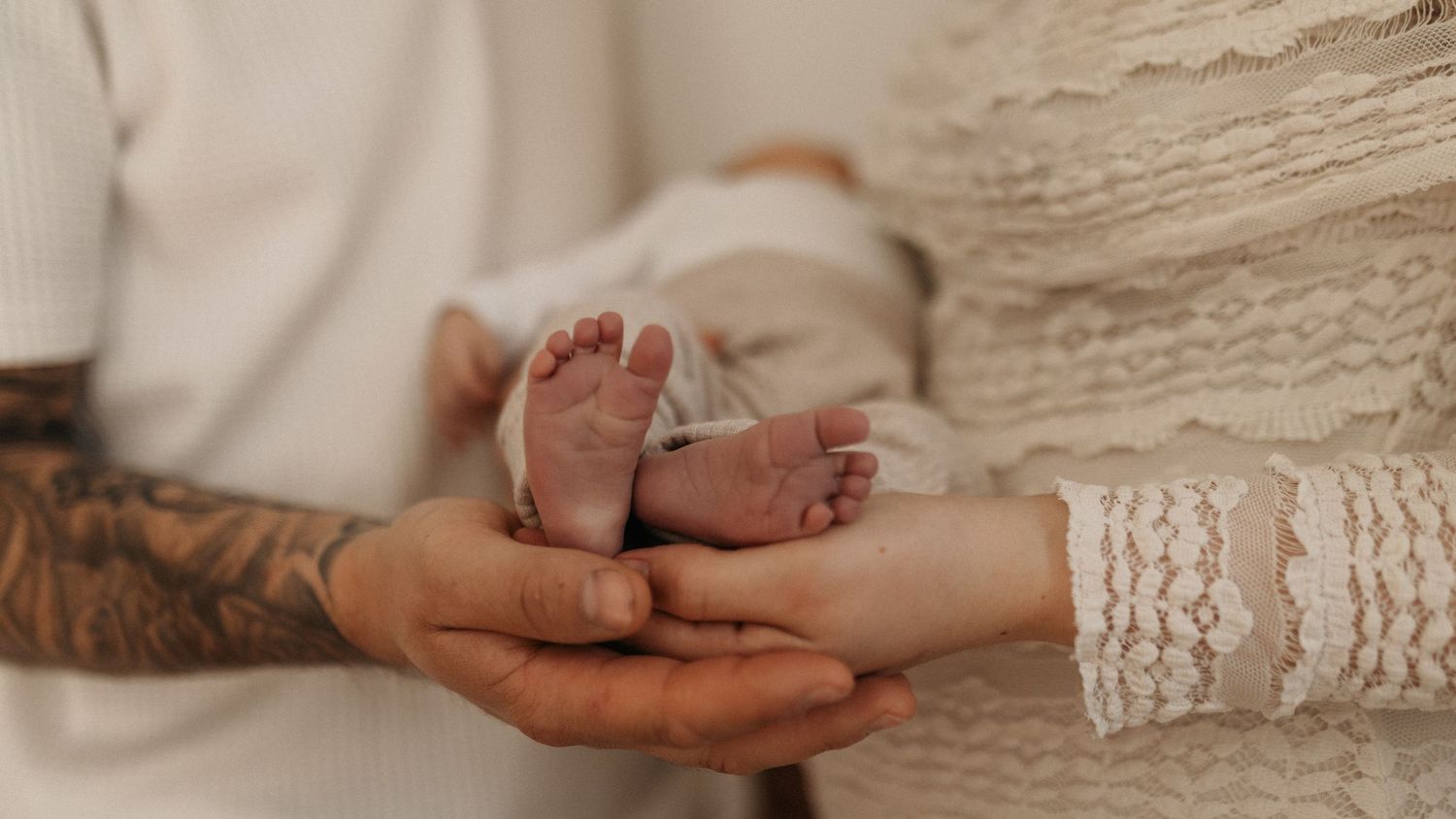 Tattooed parent and partner cradling tiny newborn baby feet in their hands, soft white background.