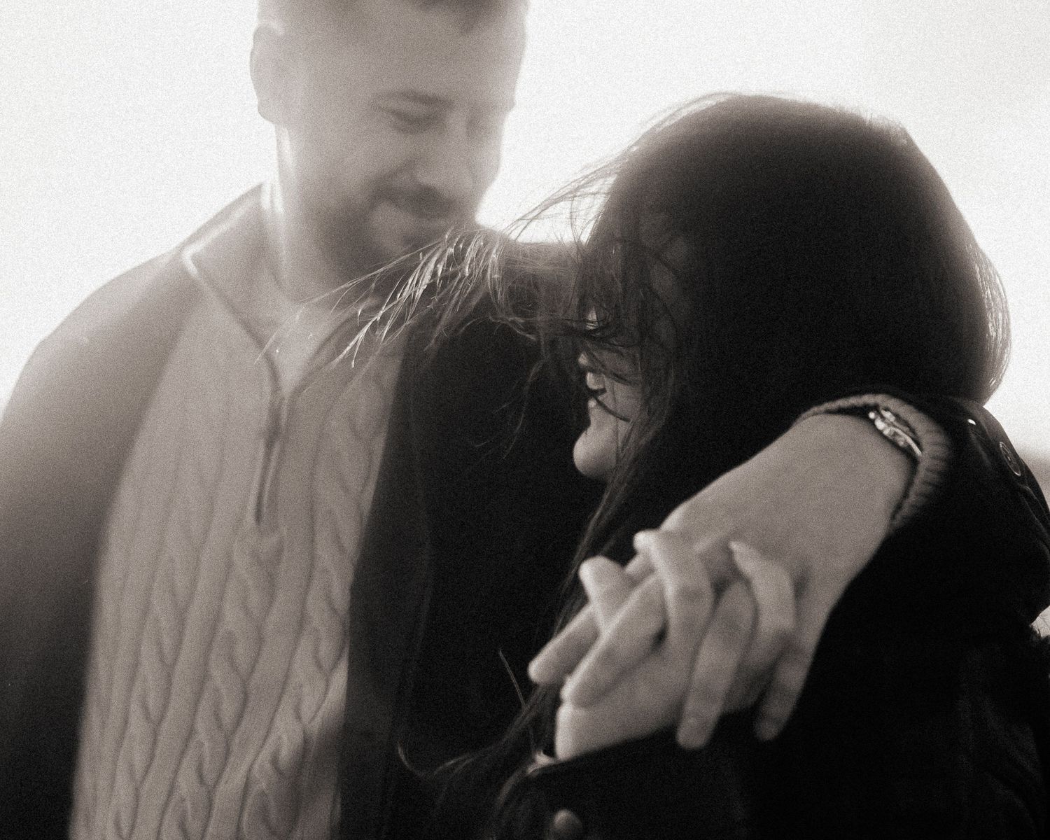 Black and white photo of a couple sharing an intimate moment, man embracing woman from behind outdoors.
