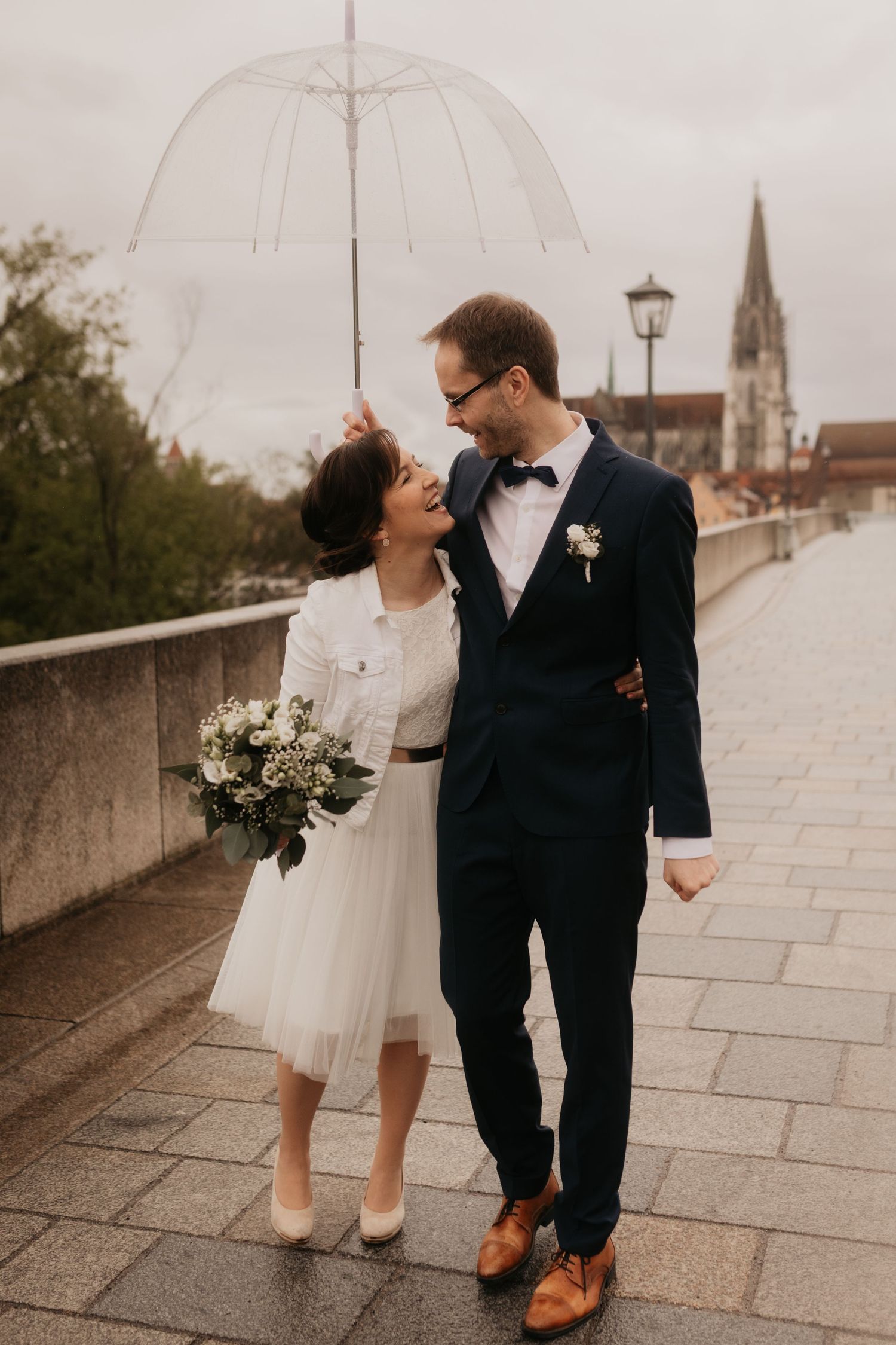 Happy newlyweds walk under clear umbrella on cobblestone bridge, bride in white dress holding bouquet, groom in navy suit.