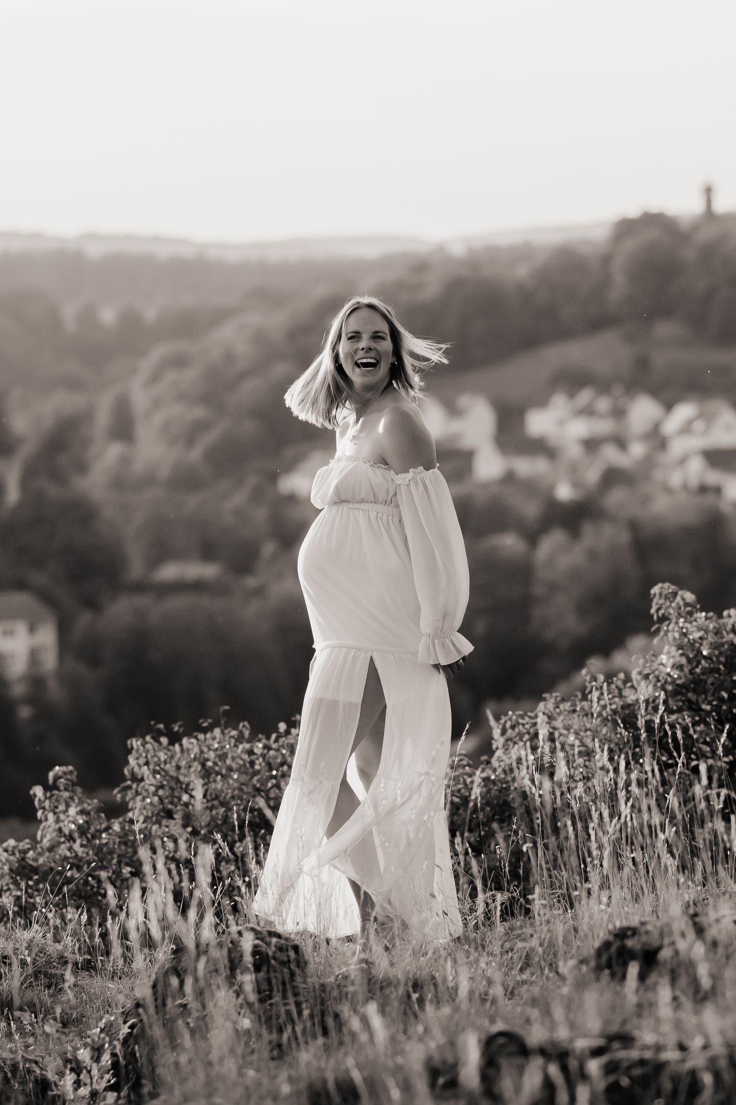 Black and white photo of a laughing pregnant woman in a white off-shoulder gown standing on a hillside.