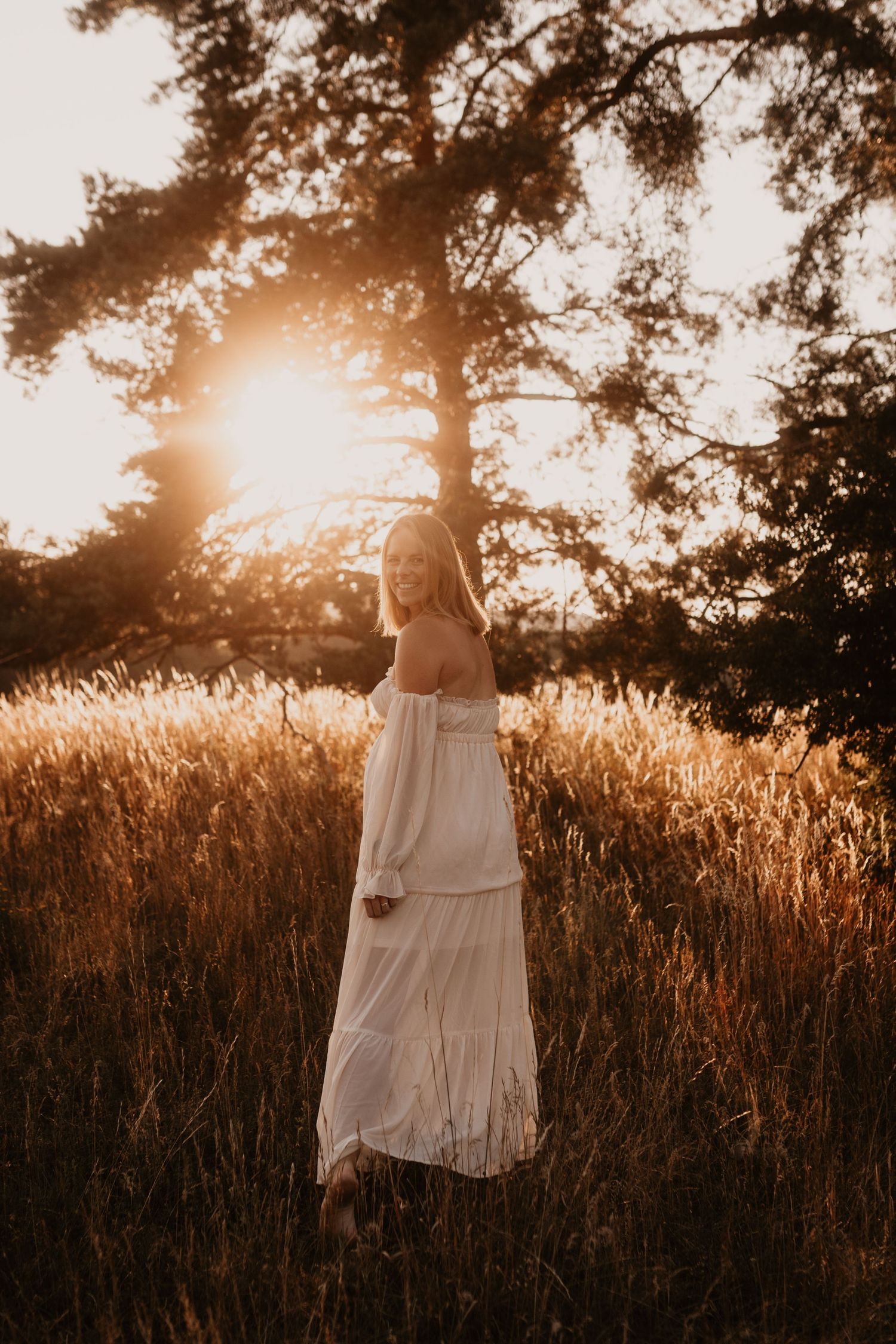 Woman in white off-shoulder dress standing in golden field at sunset with trees glowing behind her.