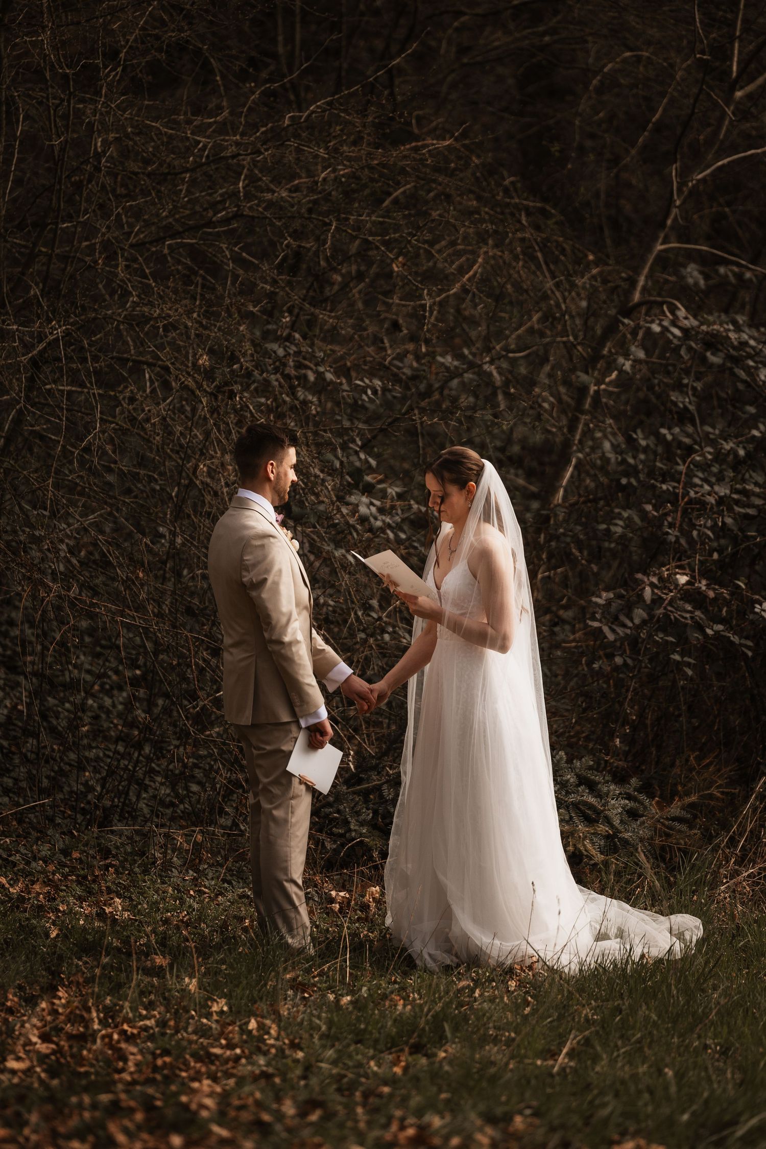 Bride and groom exchange vows outdoors in a forest, bride in white gown with veil, groom in beige suit.