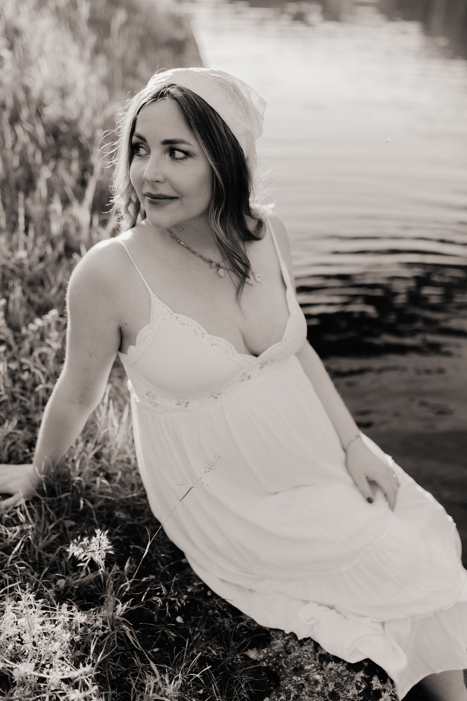 Black and white photo of a woman in a white dress and headscarf sitting by a serene lake shore.