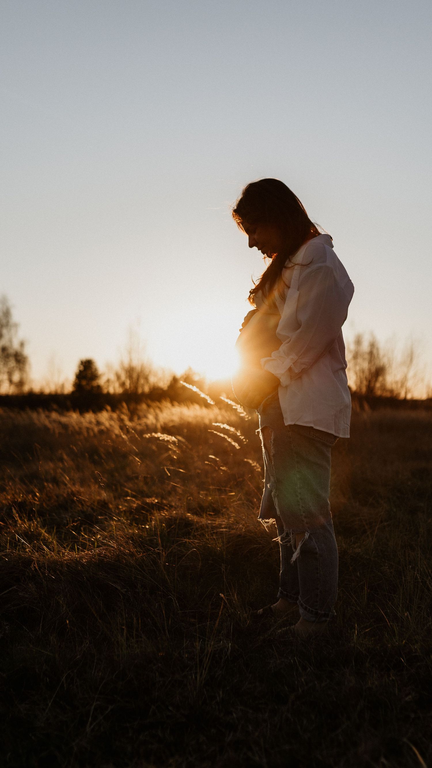 Pregnant woman cradling her baby bump in a golden sunset field, backlit by warm sunlight.