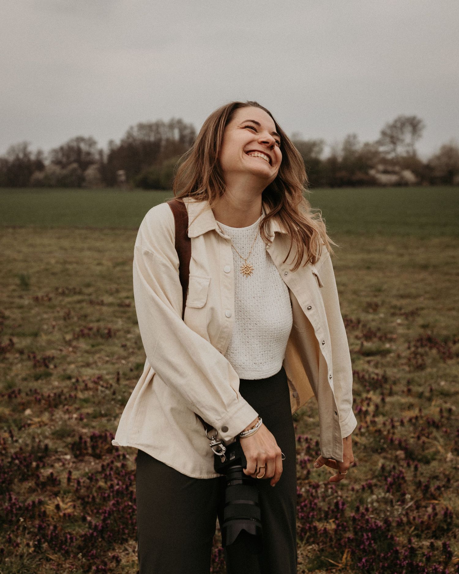 A smiling woman in a cream jacket and white top stands in a green field, looking up joyfully on a cloudy day.