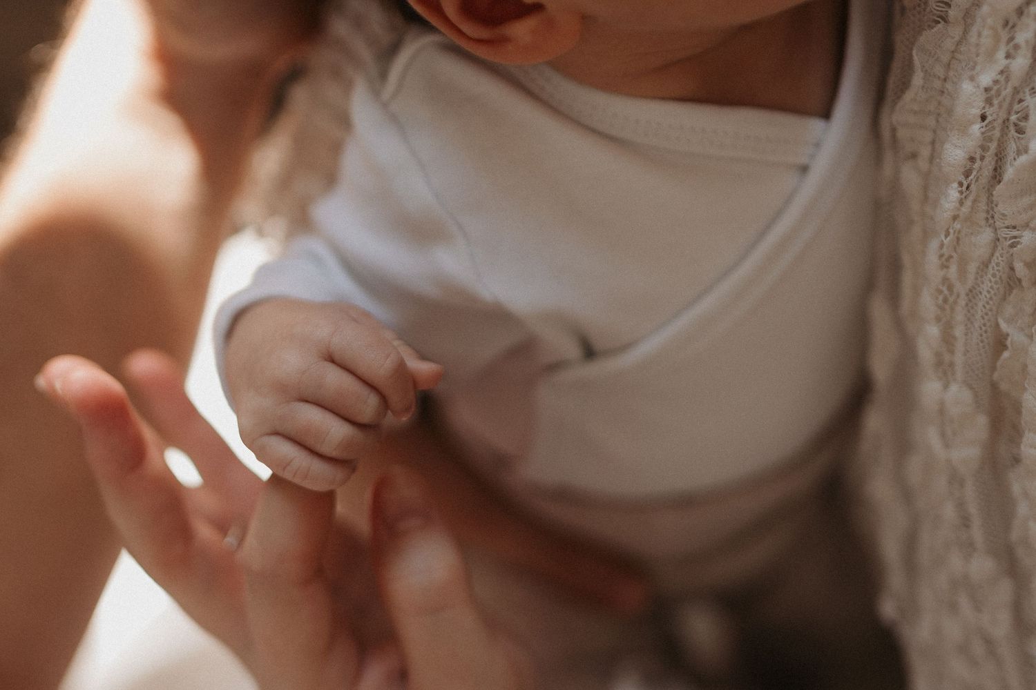 Close-up of a newborn baby grasping an adult's finger, wearing a white onesie in soft warm light.