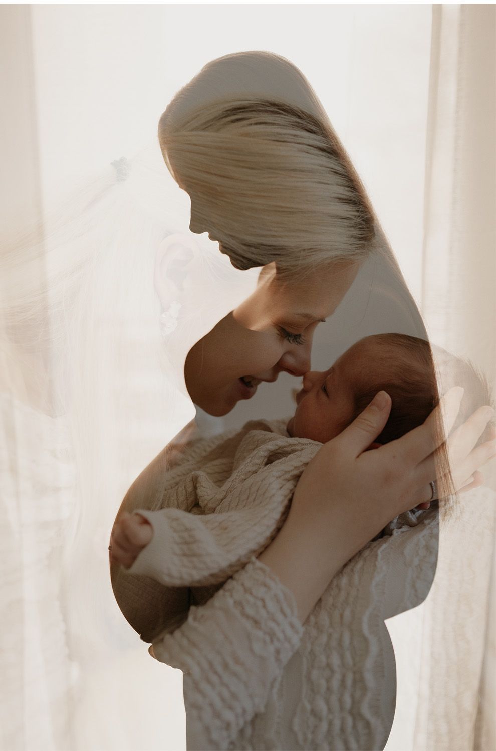 A tender moment as a blonde mother gently touches noses with her newborn baby wrapped in a soft cream knit blanket.