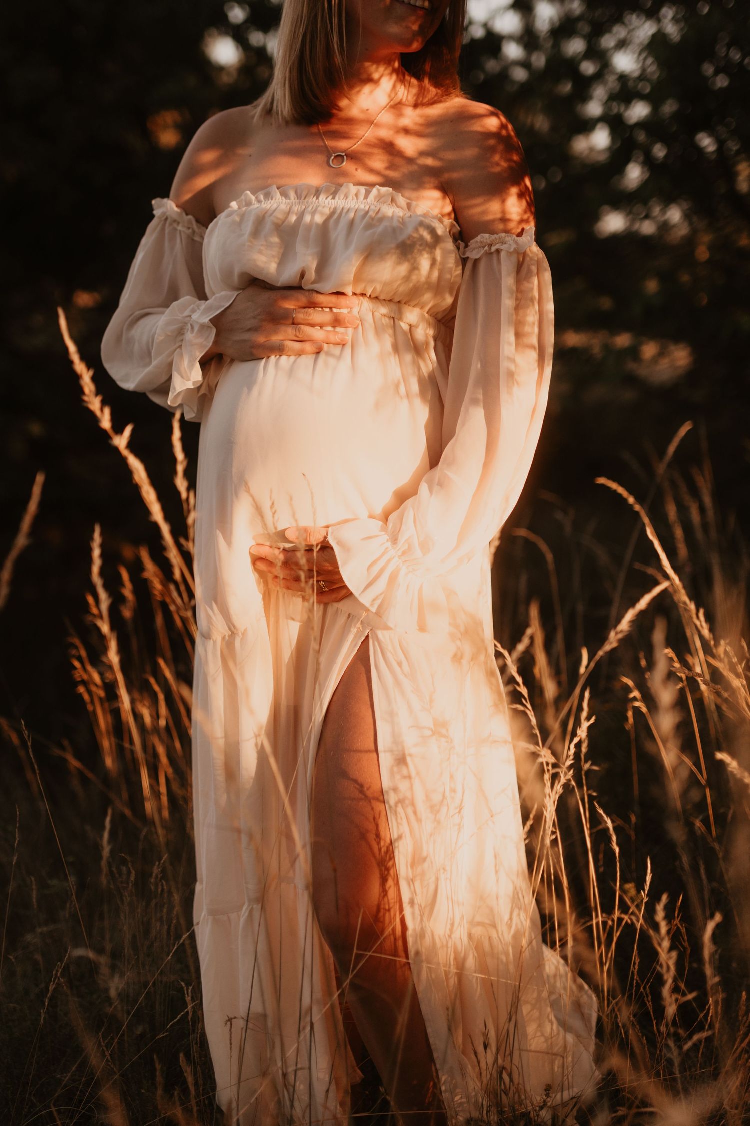 Pregnant woman in flowing white off-shoulder dress standing in golden sunlit field, cradling baby bump at sunset.