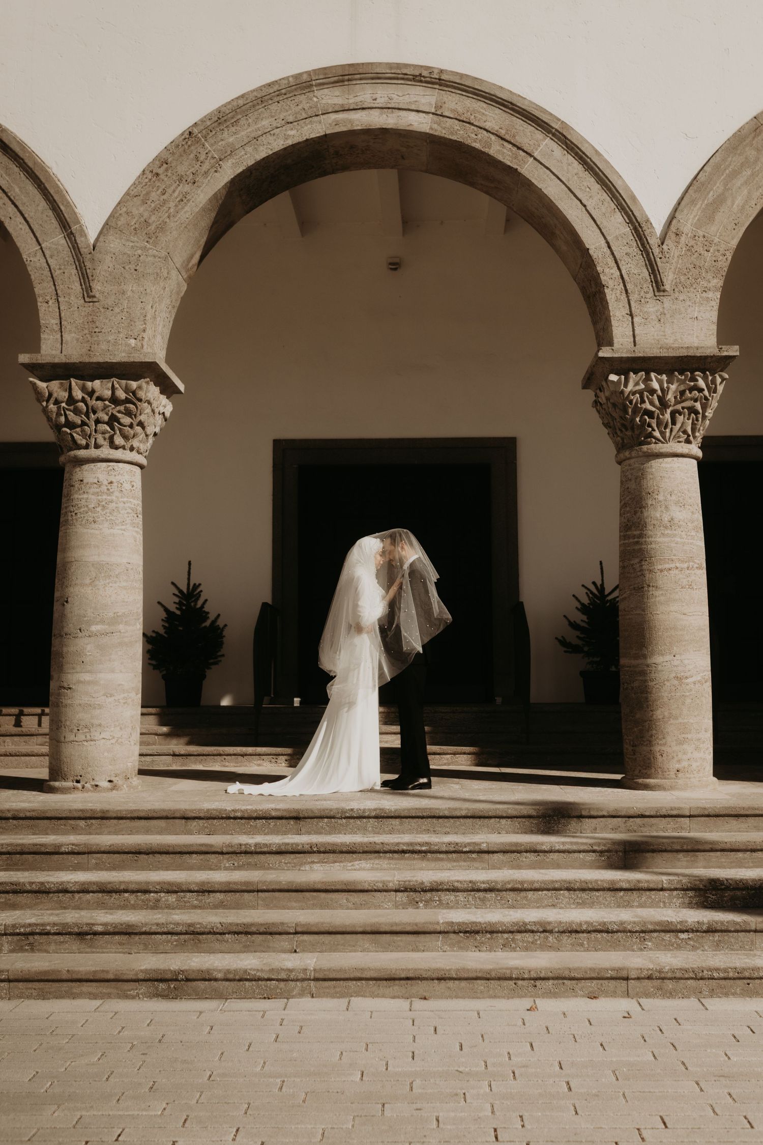 Bride and groom kissing under stone arches at a classic venue, bride wearing a flowing white gown and veil.