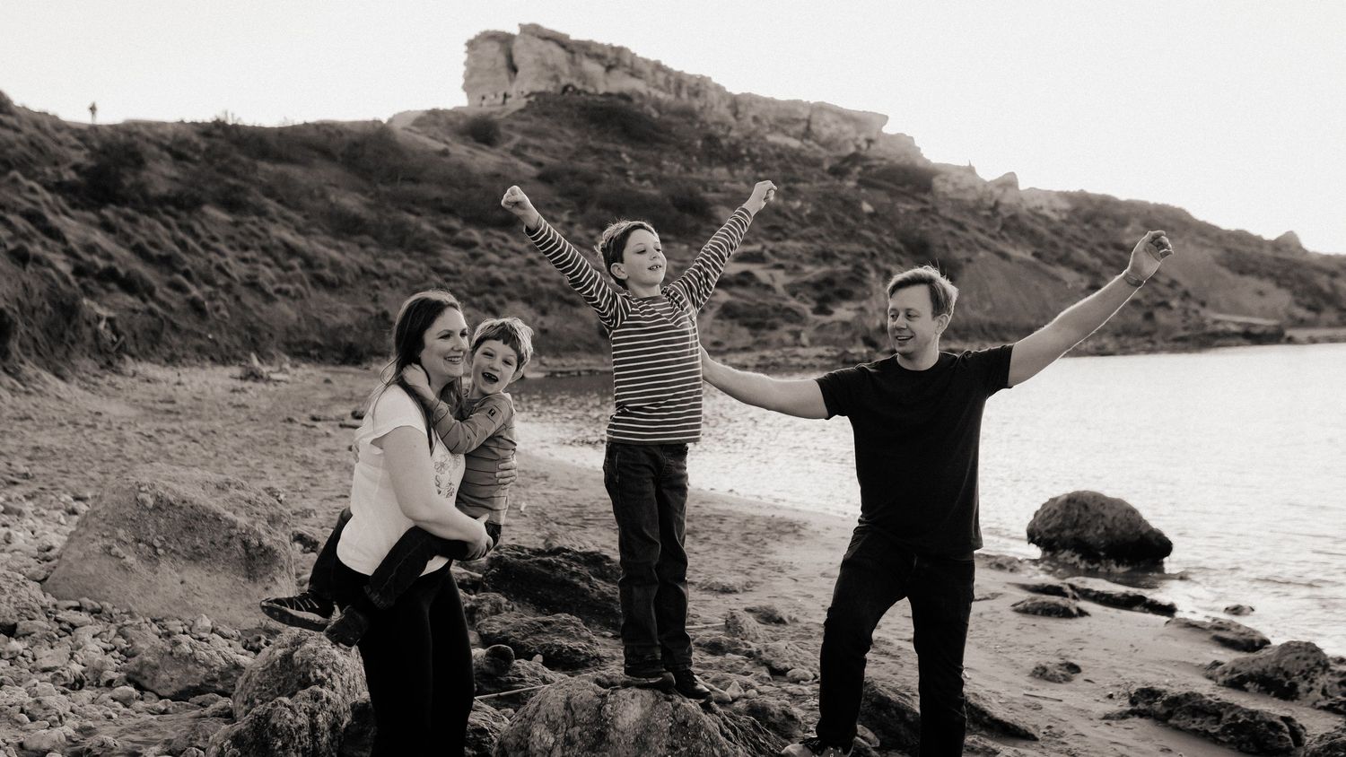 Black and white photo of a happy family of four posing on a rocky beach with a historic clifftop fortress in the background.