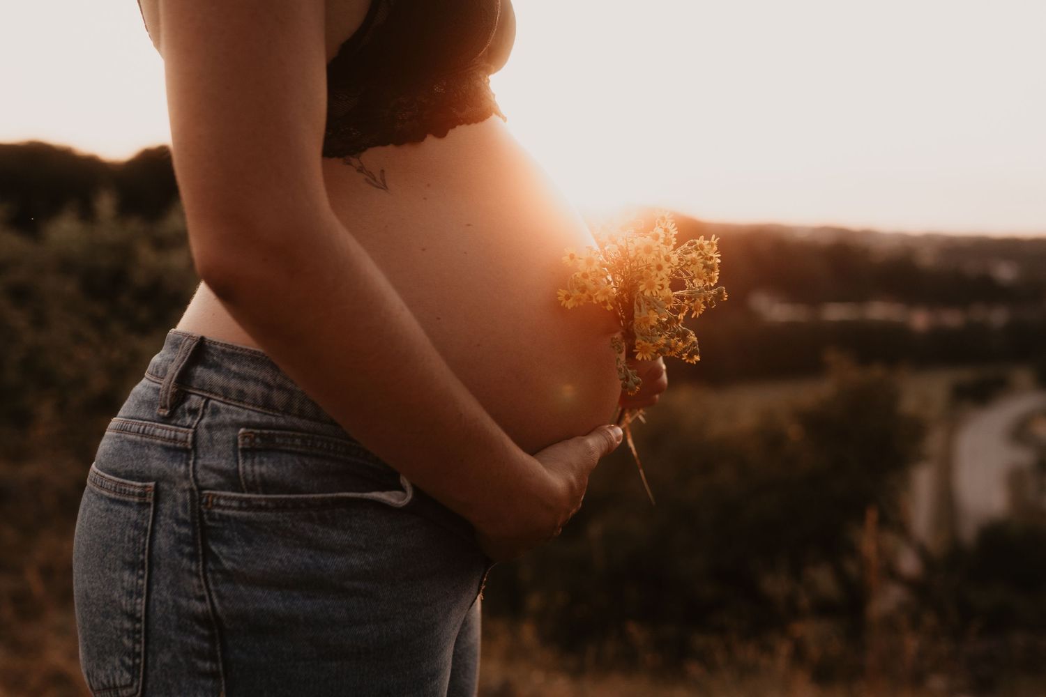 Pregnant woman in jeans holding wildflowers at golden sunset, showcasing baby bump in serene outdoor setting.