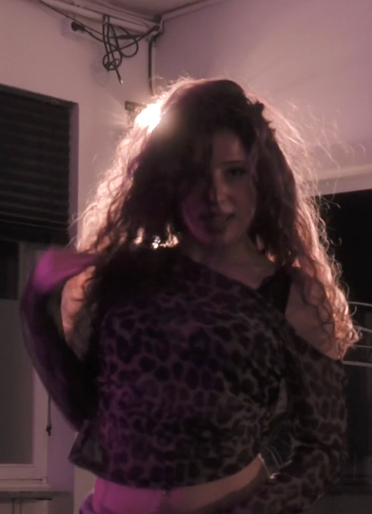 A person with long curly hair poses in a leopard print top with pink lighting in a dimly lit room.