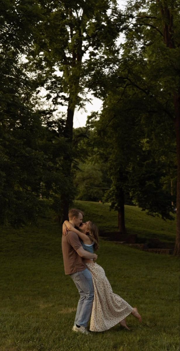A couple shares a romantic embrace and kiss in a lush green park surrounded by tall trees at dusk.