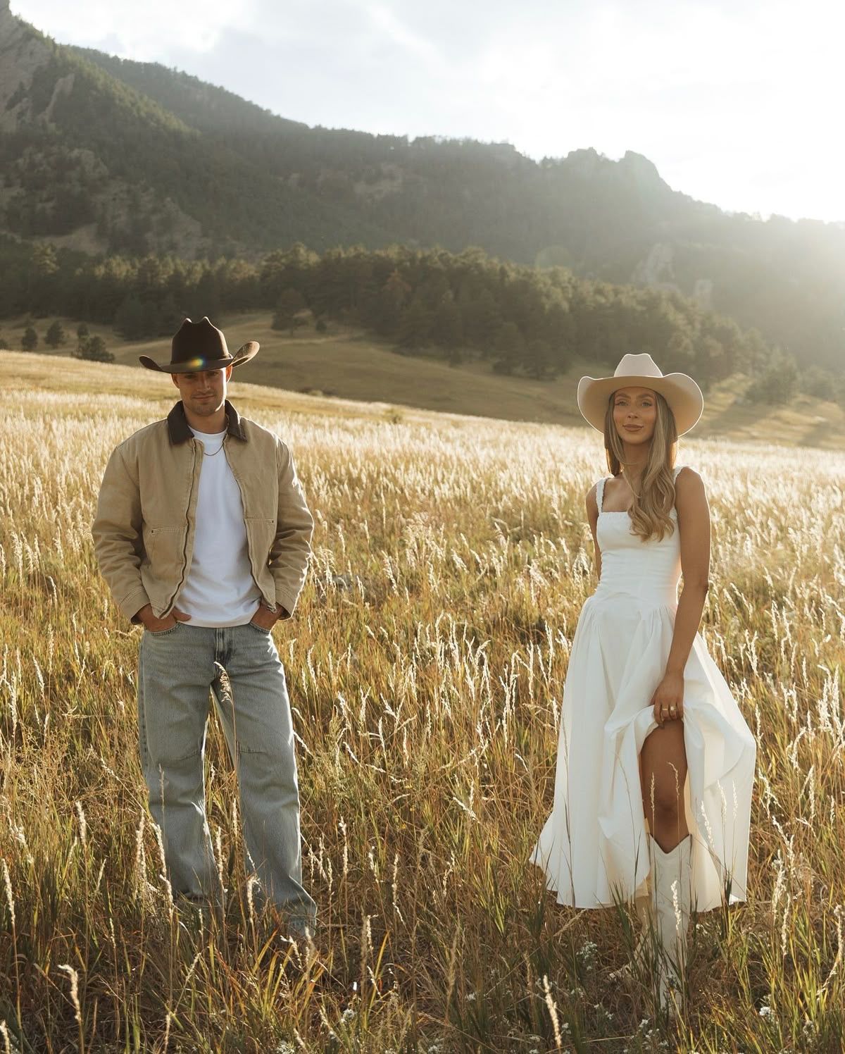 A man in a cowboy hat and a woman in a white dress stand in a golden field with mountains behind them.