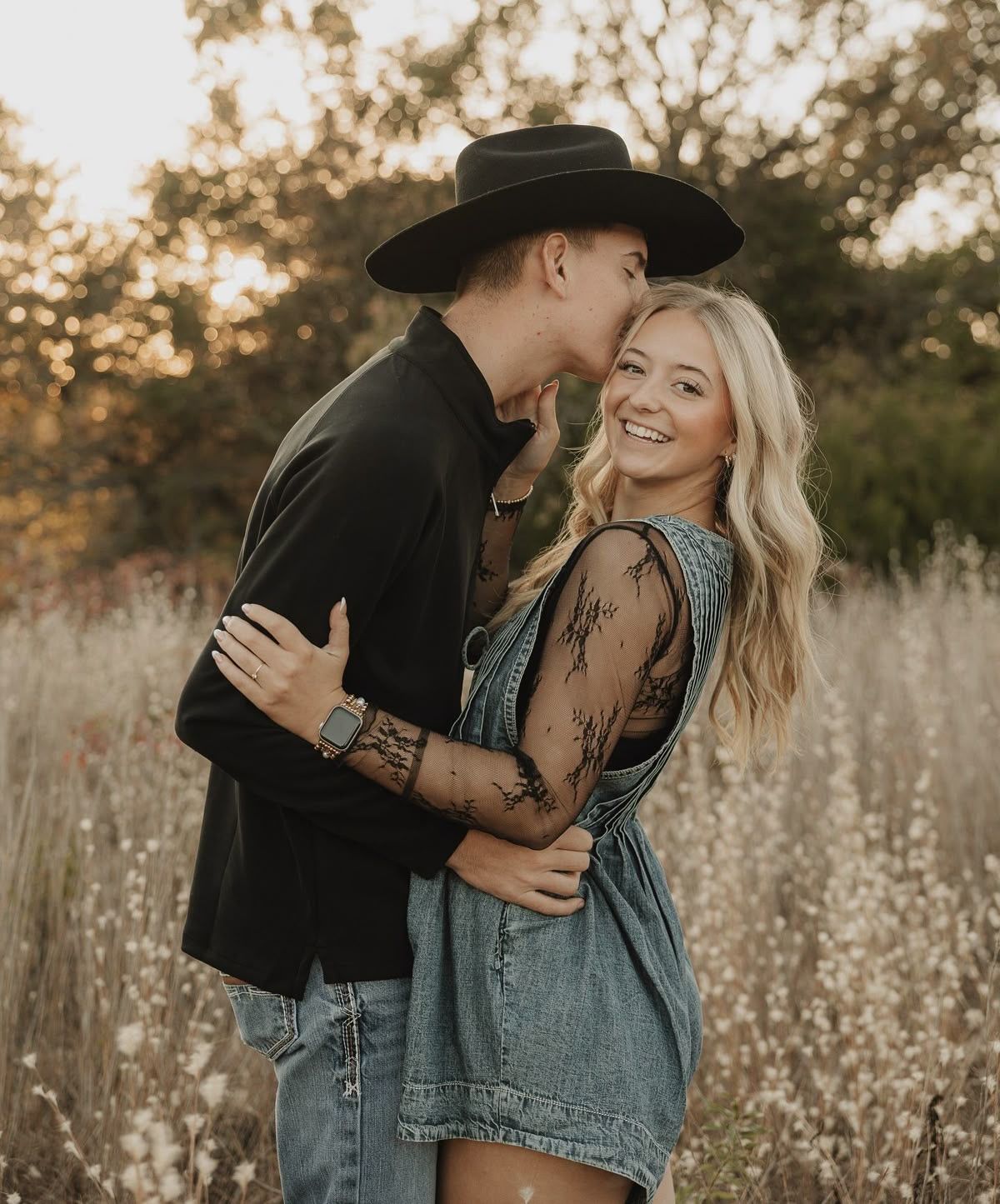 A couple embraces in a golden field, man in black hat kissing womans head as she smiles, wearing denim dress.