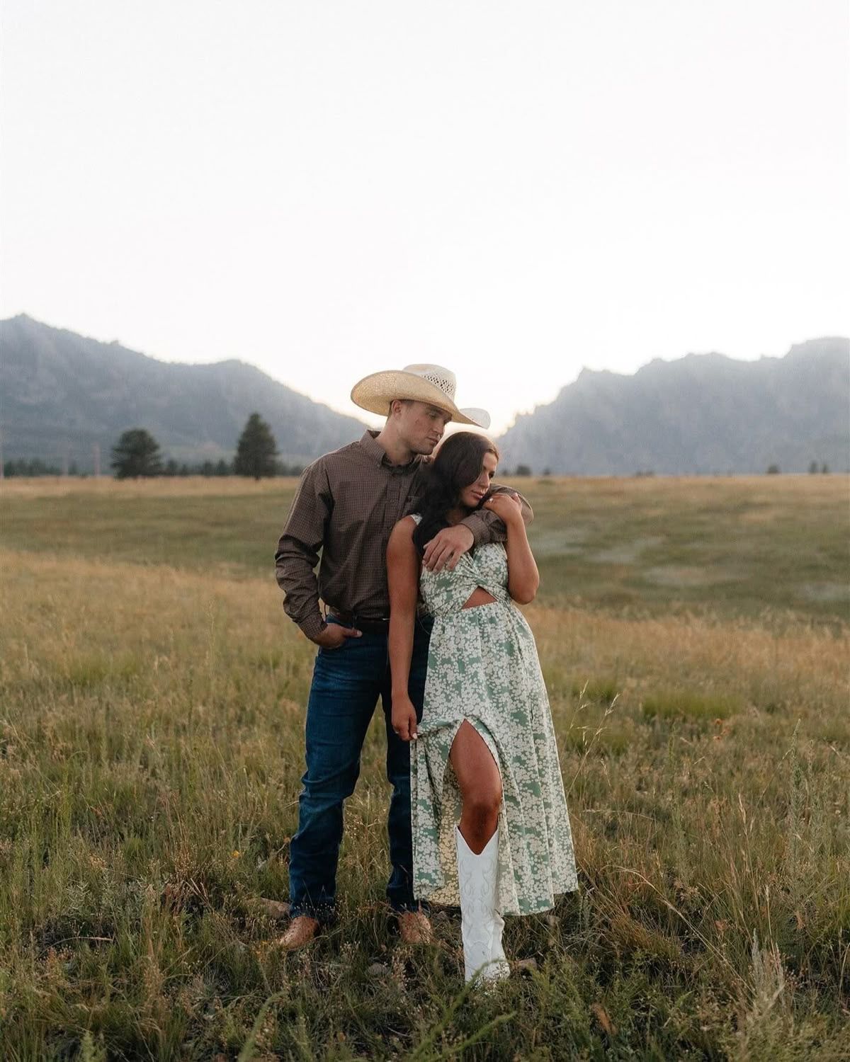 Couple embracing in a mountain meadow, man in cowboy hat kissing woman in floral dress and white boots.