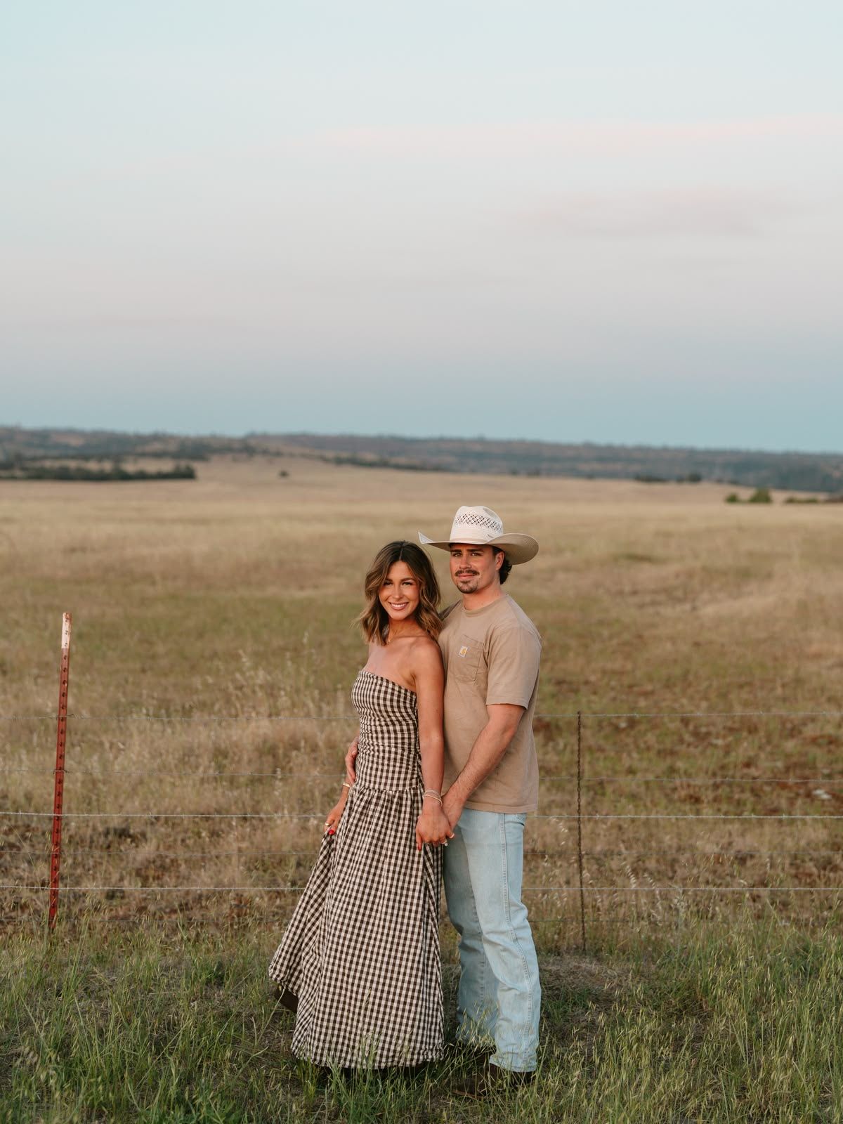 Couple holding hands in a golden field at dusk, woman in gingham dress, man in cowboy hat, rustic fence behind them.
