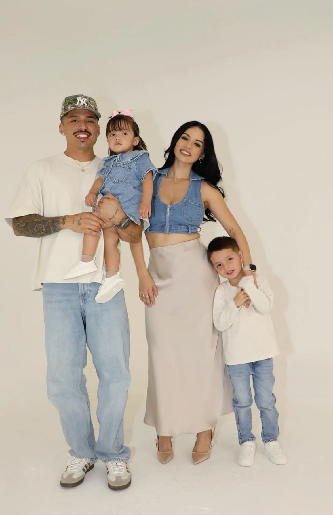 A stylish young family of four poses together in coordinated outfits against a white background.