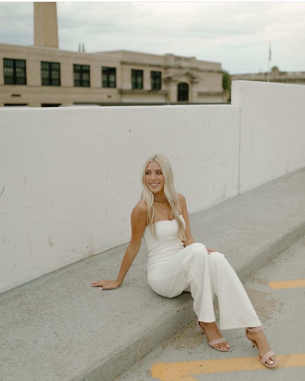 Woman in elegant white jumpsuit sitting on concrete curb with industrial building in background.