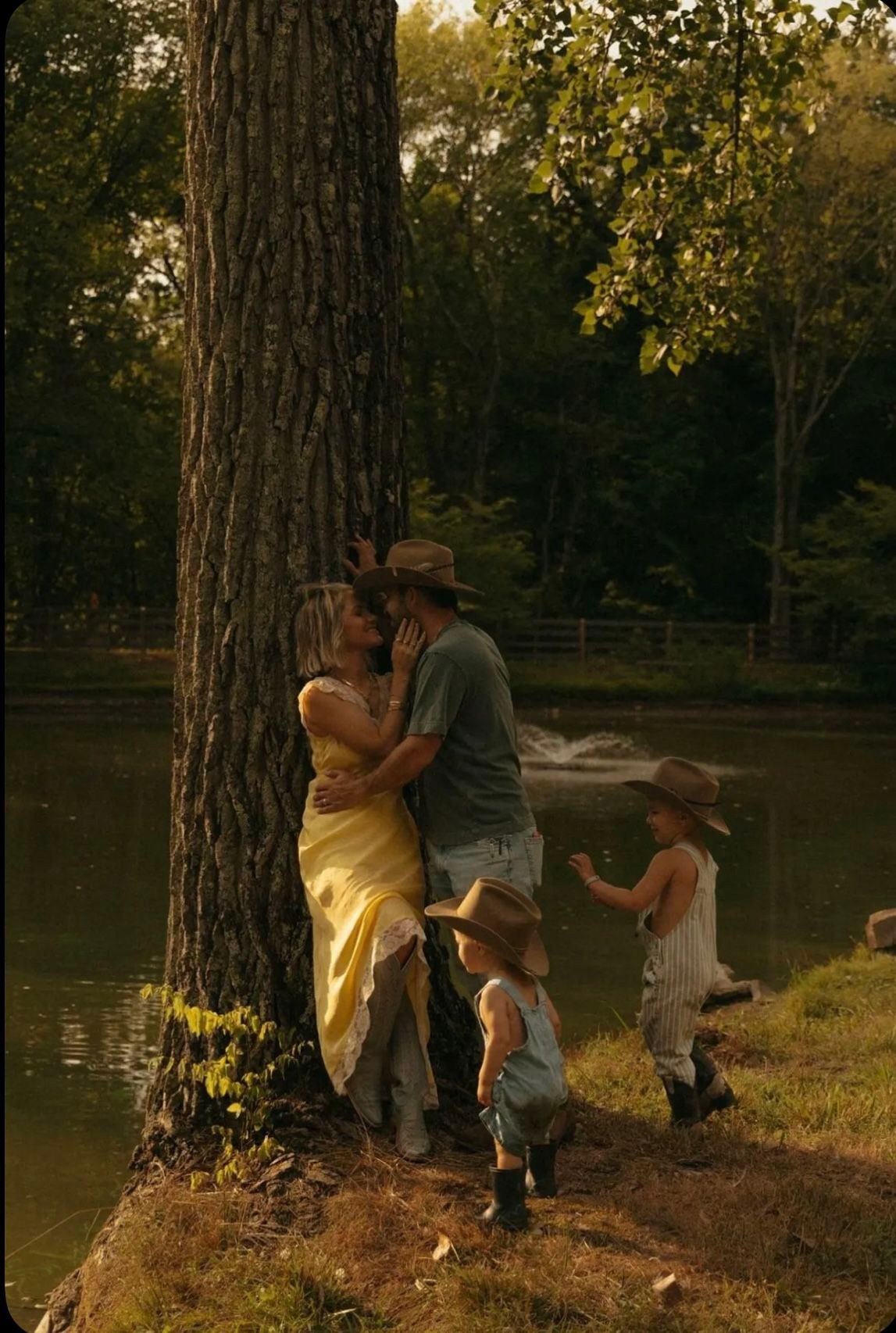A family shares tender moments by a tree near a tranquil lake during golden hour, creating a warm and intimate scene.