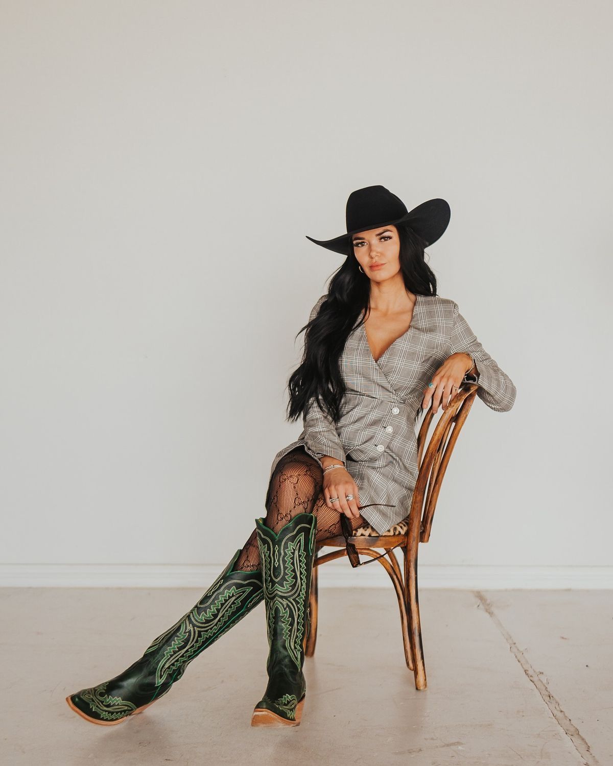Woman in black cowboy hat, plaid dress, fishnet tights, and green cowboy boots seated on a wooden chair.