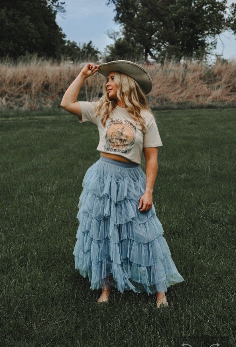 Woman in blue ruffled tulle maxi skirt, graphic crop tee, and wide-brim hat posing outdoors on green grass.