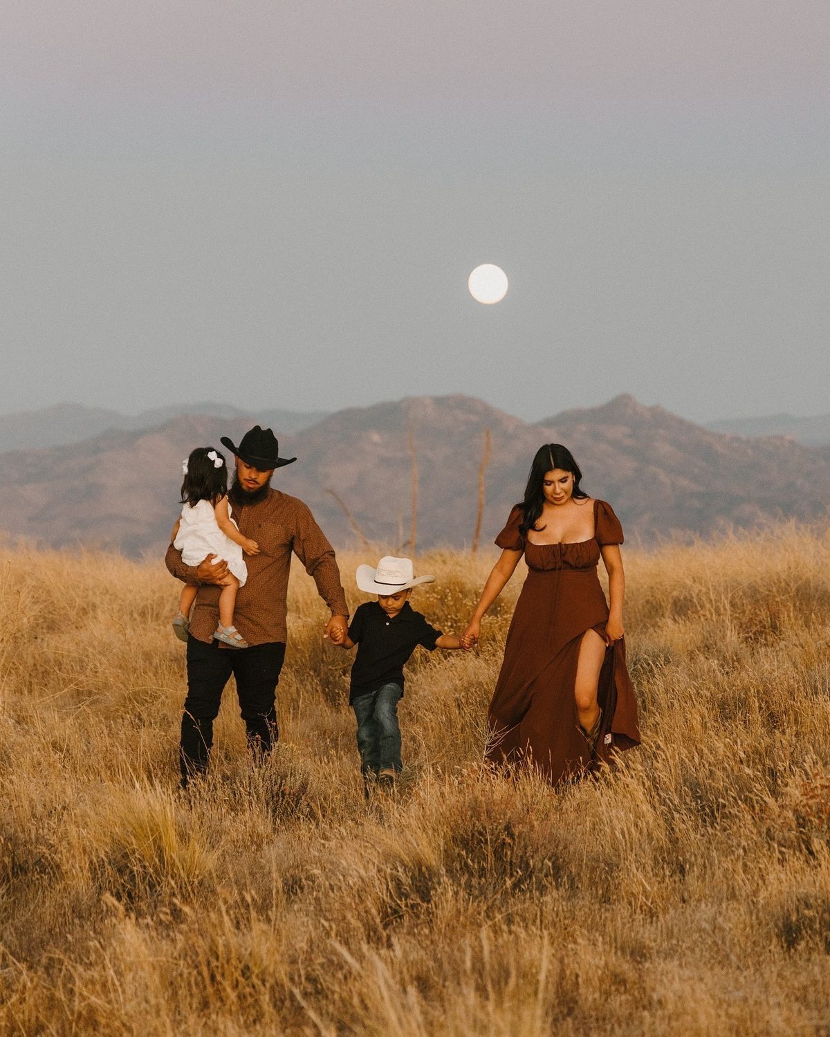 Family of four in western attire walking through golden fields under a full moon with mountain backdrop.