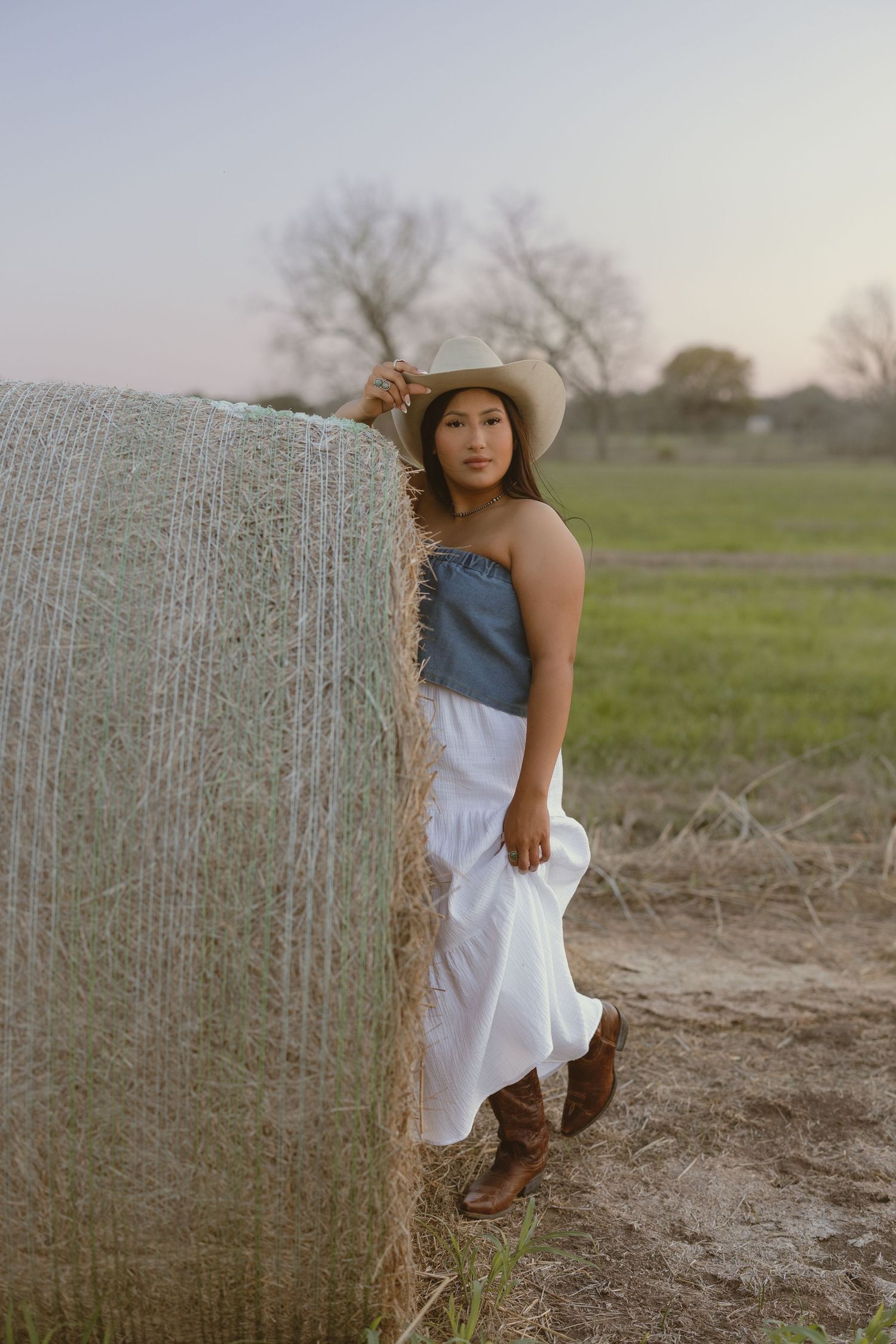A rustic country portrait featuring a straw hat, denim top, and white skirt against a hay bale in a pastoral setting.