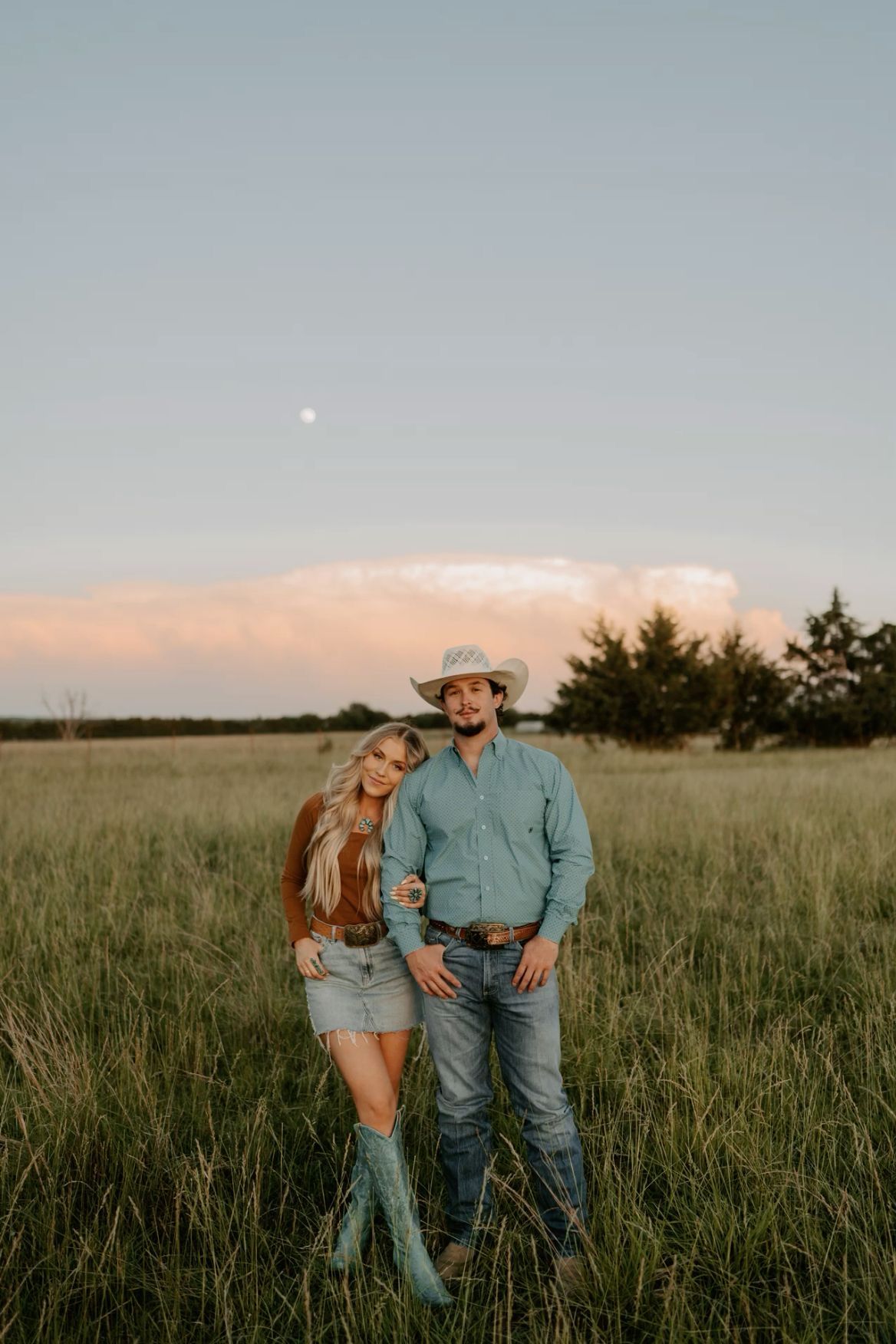 A couple in western attire poses in a grassy field at sunset, with the man in a cowboy hat and woman in turquoise boots.